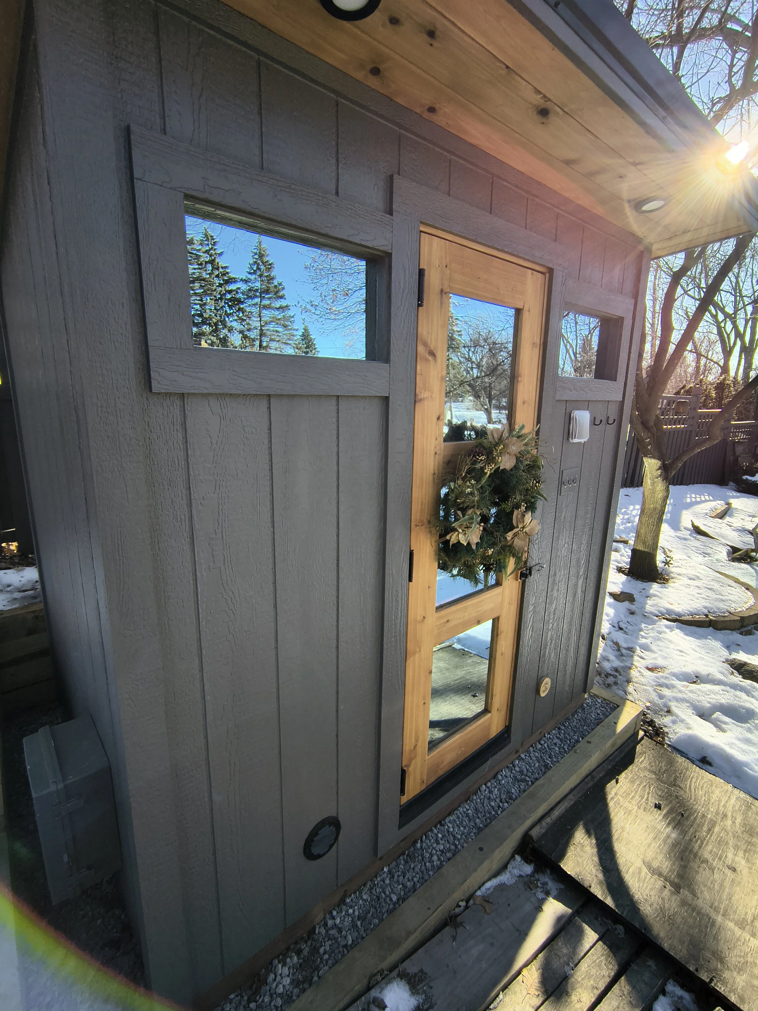 A small modern shed with gray wooden walls and a natural wood door decorated with a holiday wreath, placed outdoors in snow on a sunny day.