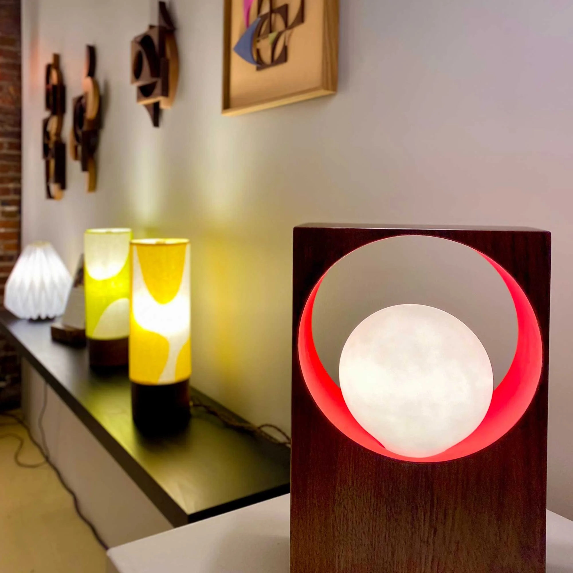 A modern interior space with decorative lamps on display. The foreground features a unique lamp with a wooden frame and glowing sphere. In the background, there are cylindrical lamps with yellow and orange patterns on a black shelf, along with framed wall art and wooden wall sculptures.