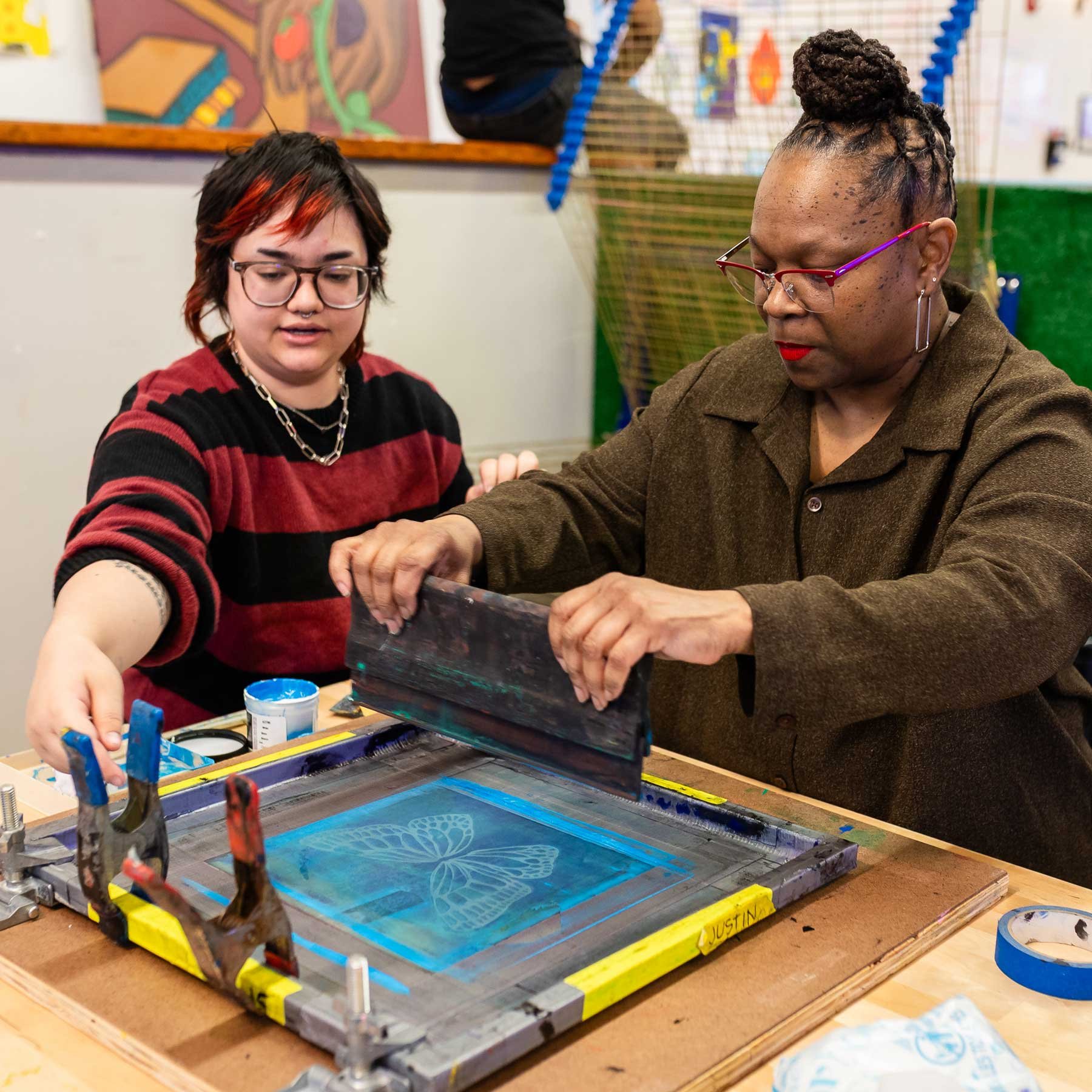 Two people engaged in screen printing art on a table, with various tools and supplies around them.