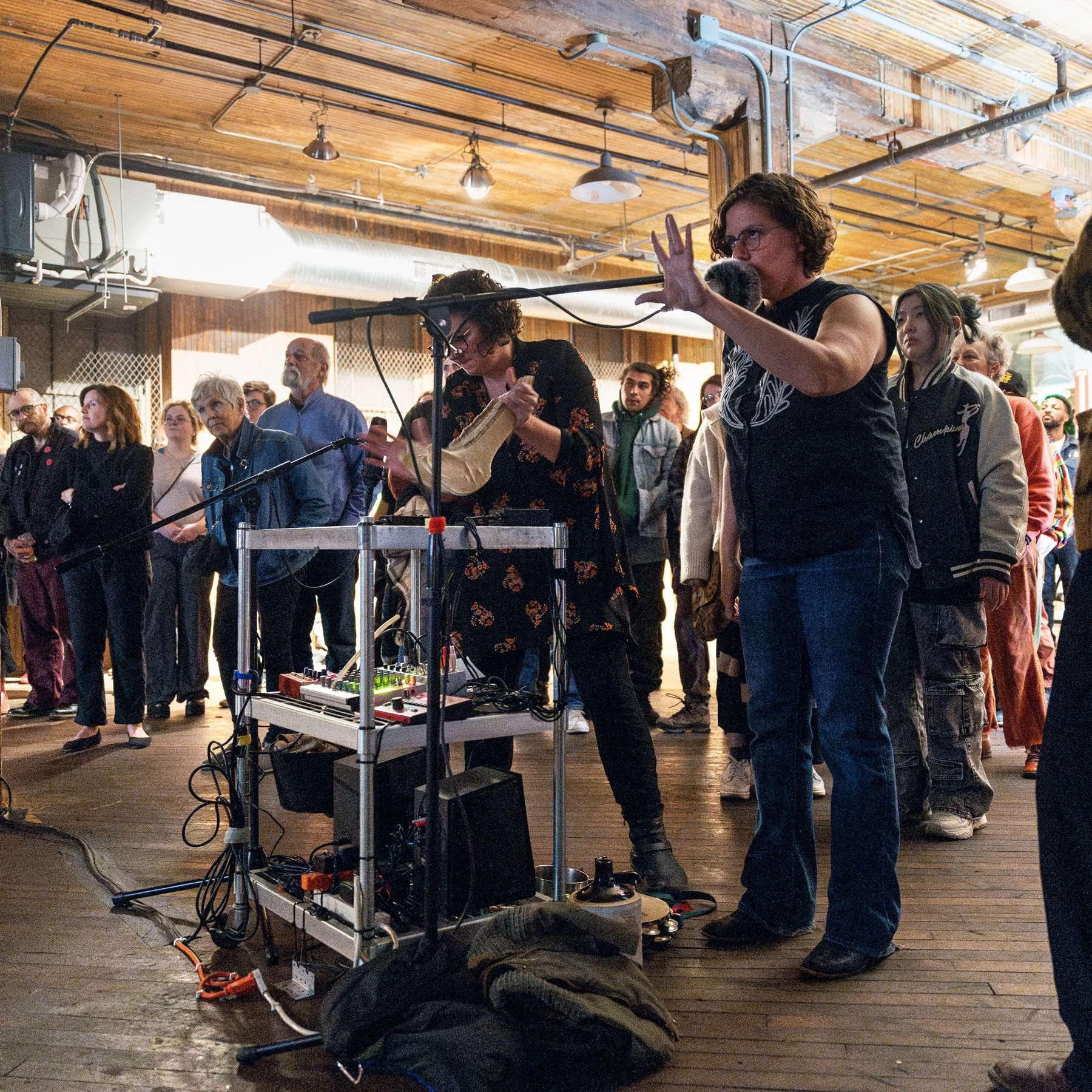 People gathered at an indoor event, focused on two individuals using electronic equipment, with microphones and a sound setup on a cart. The atmosphere is casual, in a venue with wooden floors and a high ceiling.