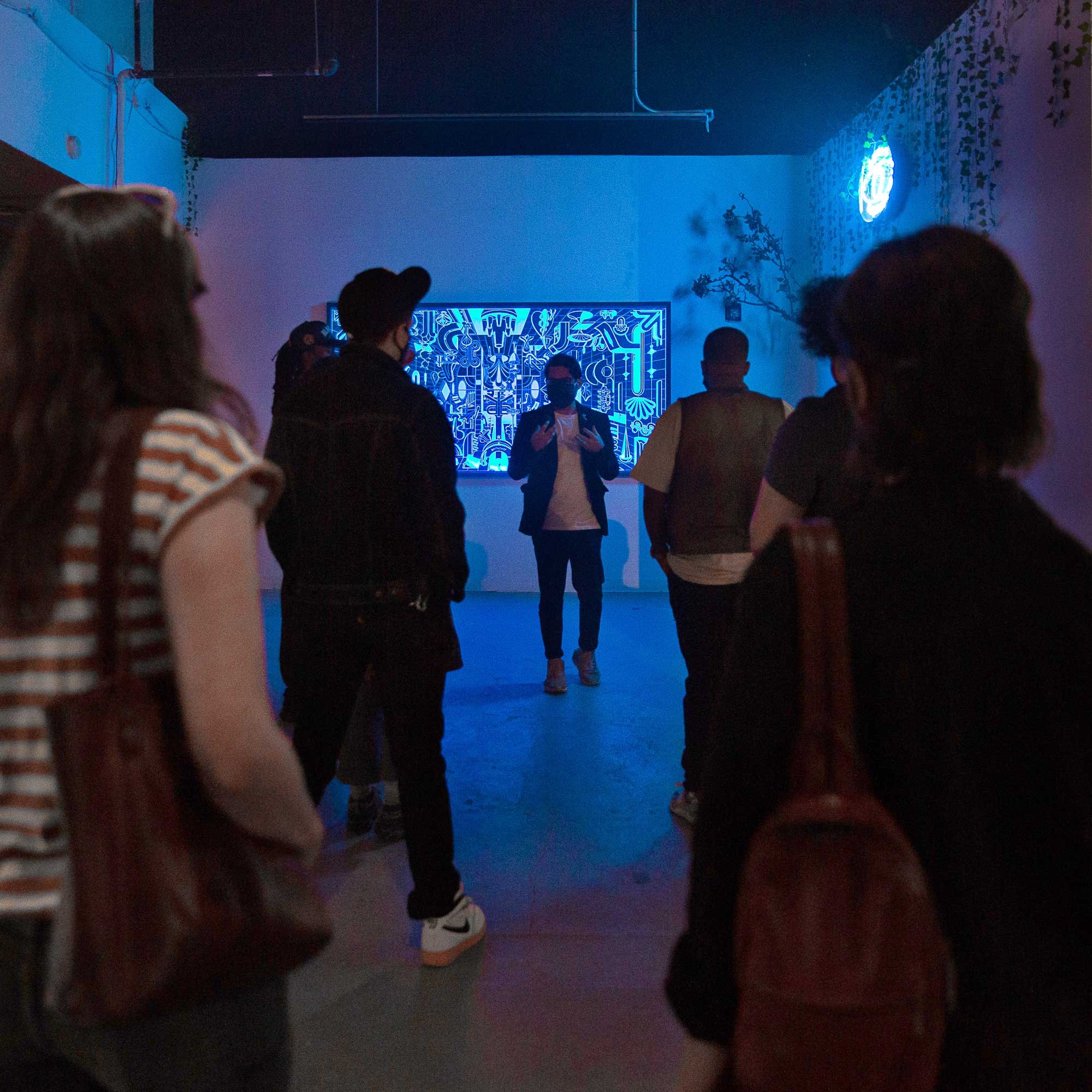 People observing a neon art display in a dimly lit gallery. The artwork features abstract blue patterns and symbols. The audience, mostly wearing casual attire, includes a person speaking near the art.