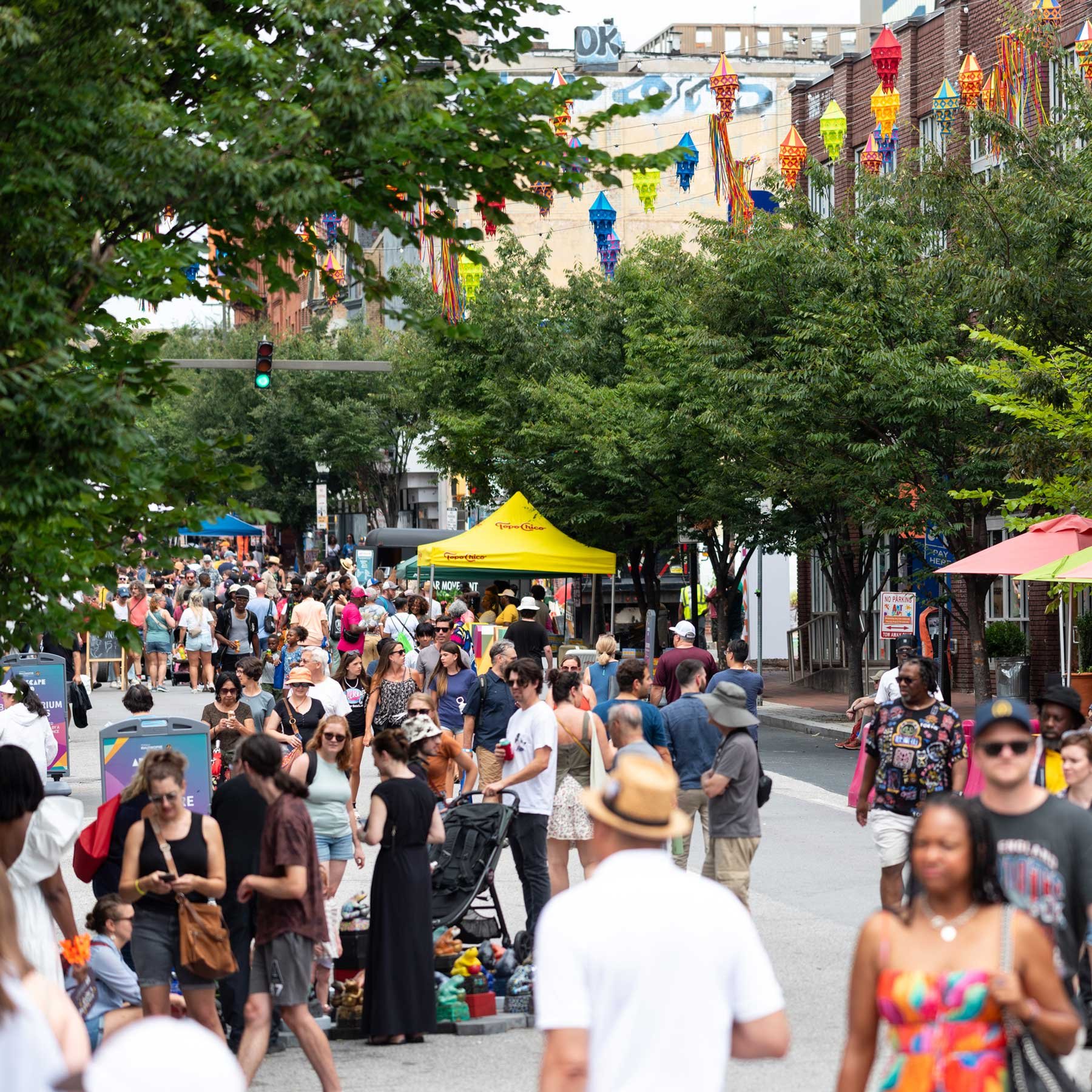 Crowded urban street festival with diverse people, colorful hanging decorations, and vendor booths.