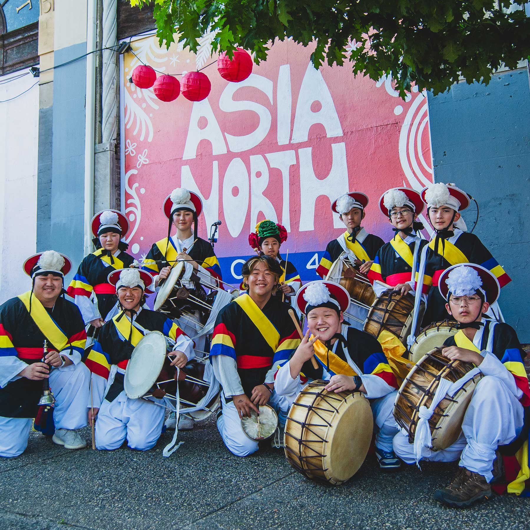 A group of people in traditional Korean costumes and hats, holding traditional musical instruments, posing in front of a wall with 'Asia North' painted on it. Red lanterns hang above them.