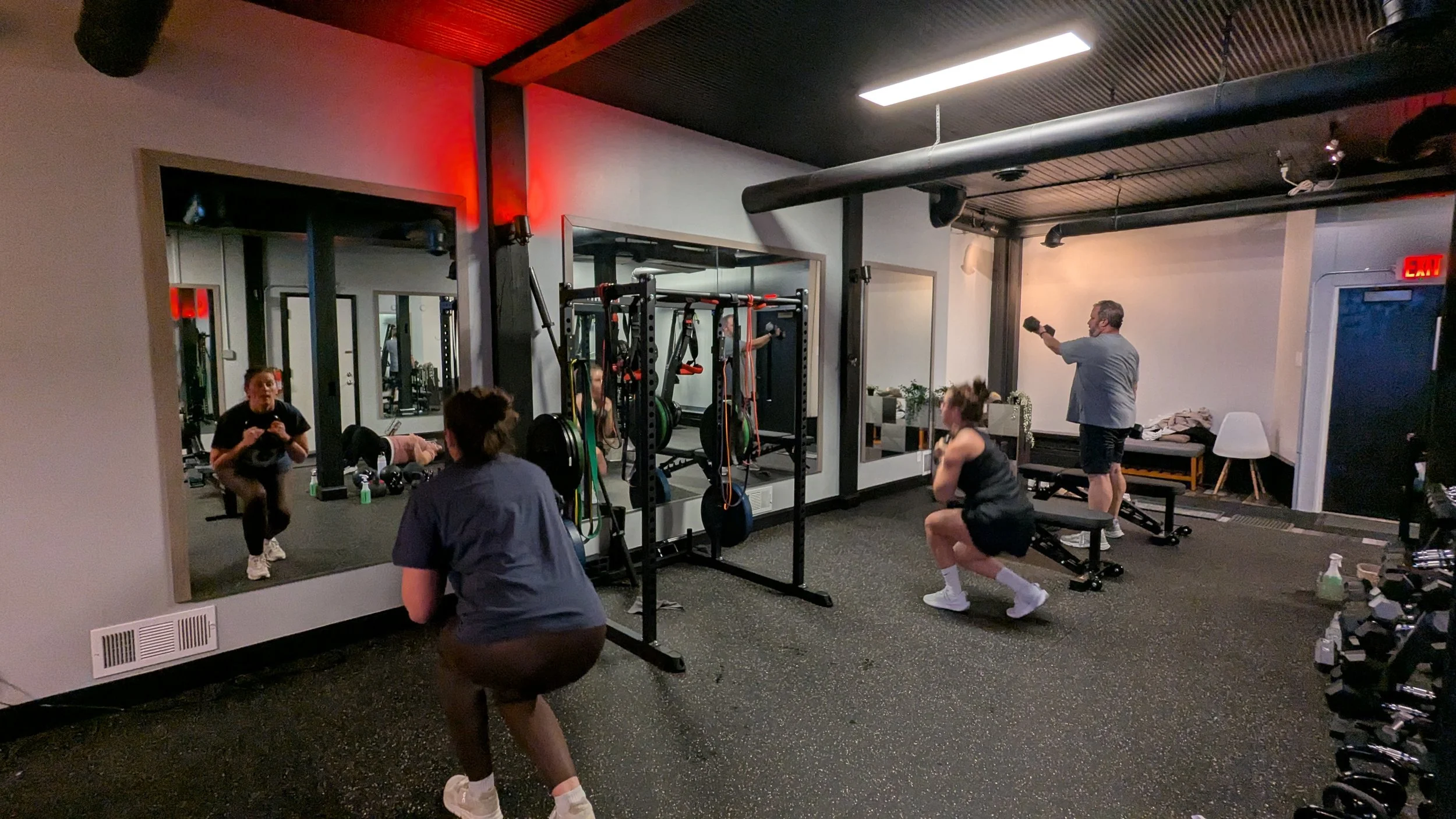 People working out in a gym, including squatting, lifting a dumbbell, and working with resistance bands, mirrored wall, and gym equipment.