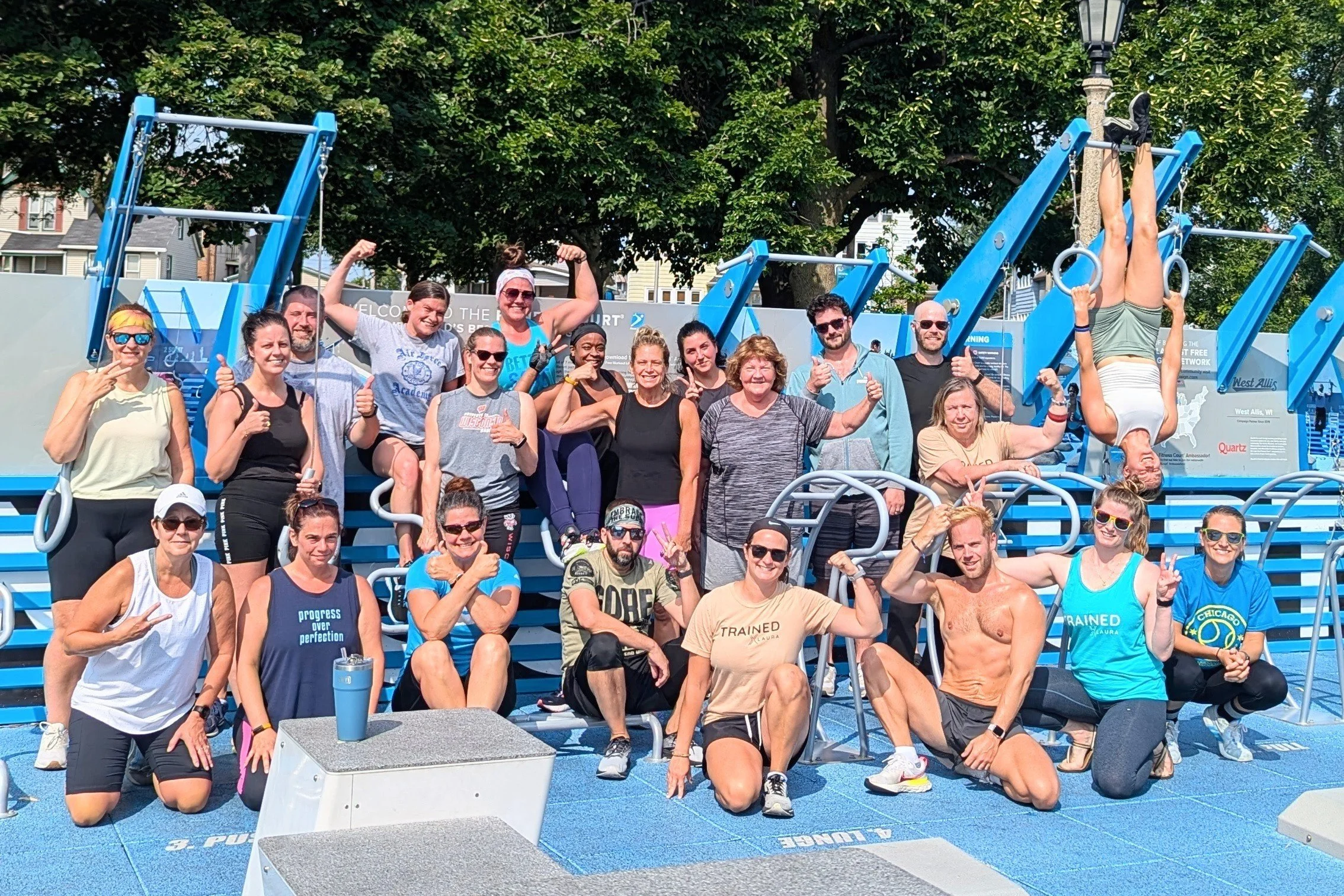 Group of people in athletic gear at an outdoor fitness park posing for a photo, with one person performing an upside-down exercise on rings.