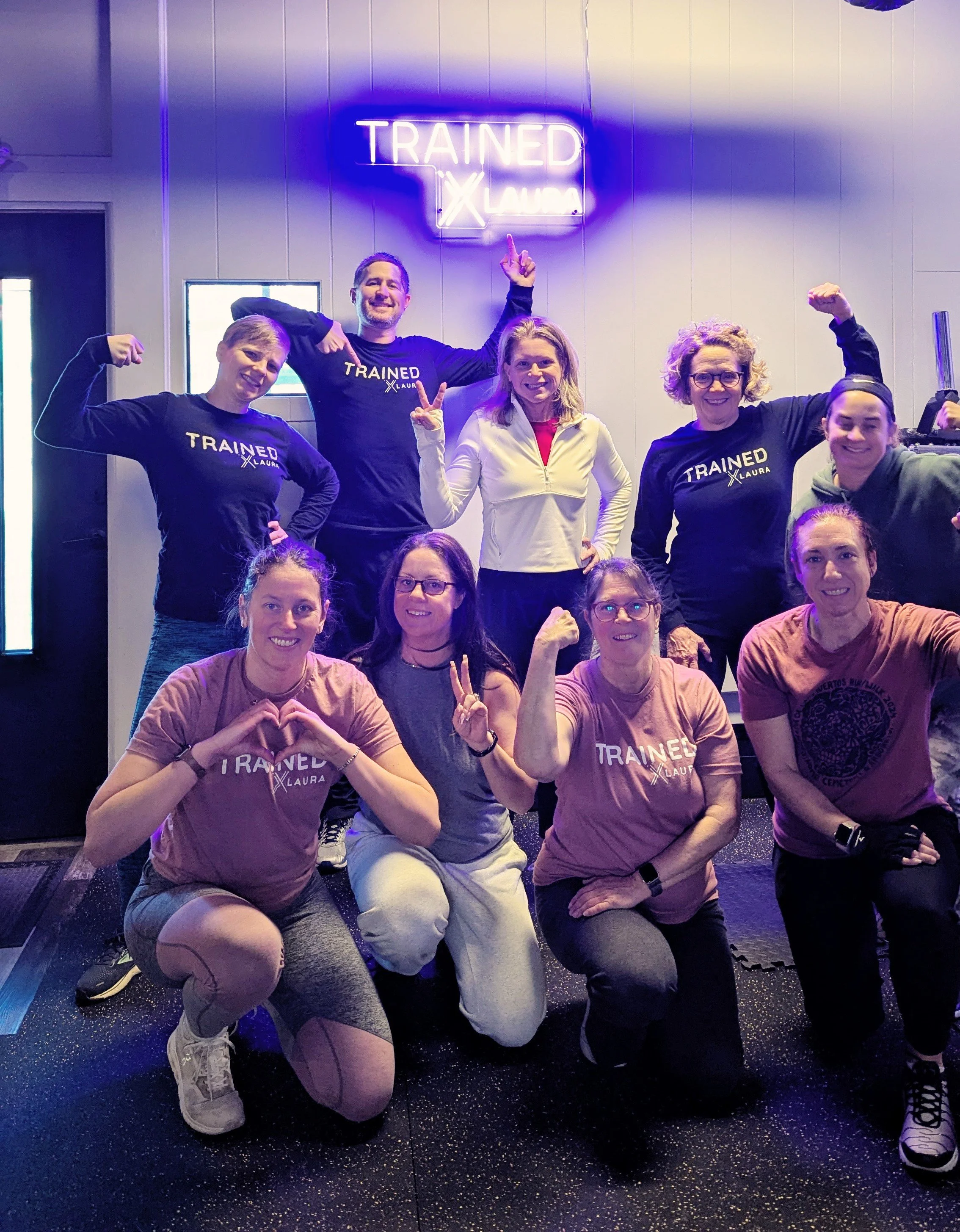 Group of women and men posing inside a gym, some wearing 'Trained by Laura' shirts, with a neon sign that reads 'Trained by Laura' in the background.