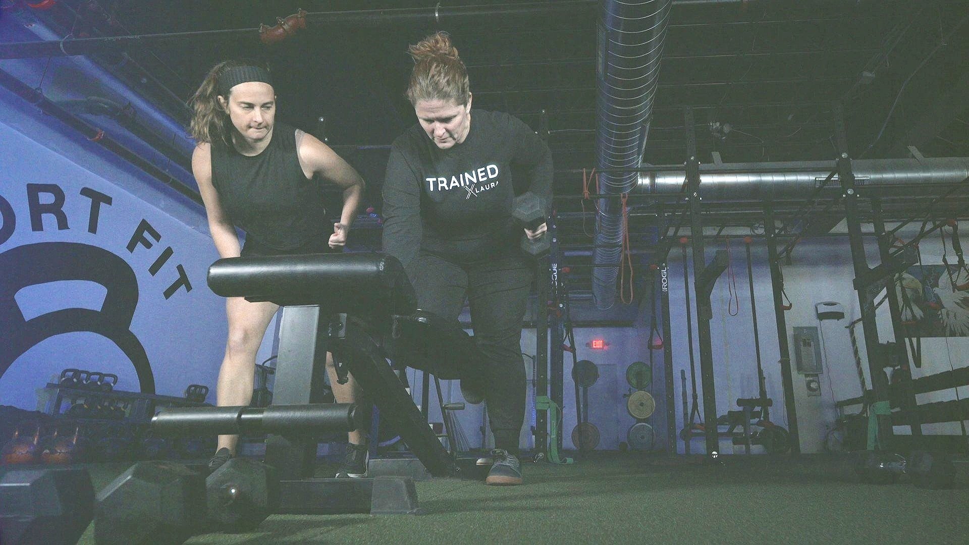 Two women working out in a gym, one with a dumbbell, both facing forward in a focused posture.