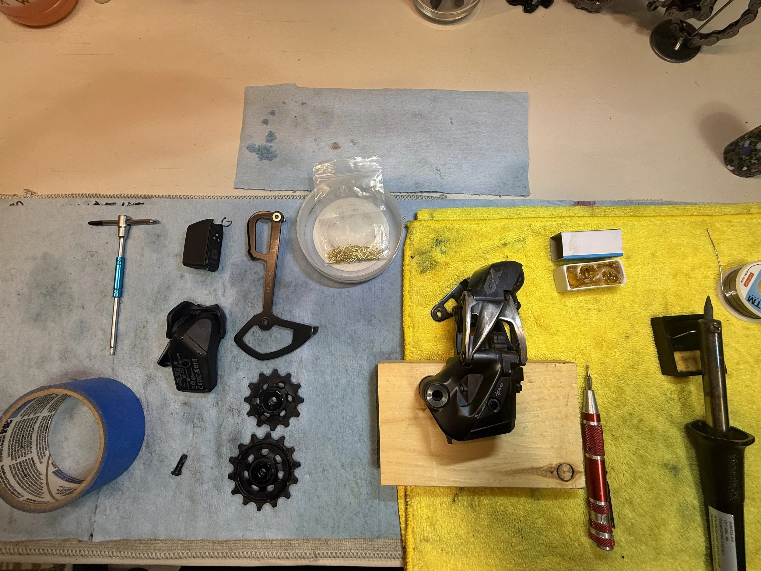 A workbench with bike repair tools and parts, including sprockets, a crank, a chainring, a crankset, a tire lever, a chainring bolt, a chainring nut, some screws, and a Shimano chainstay with tools on a yellow and gray cloth.