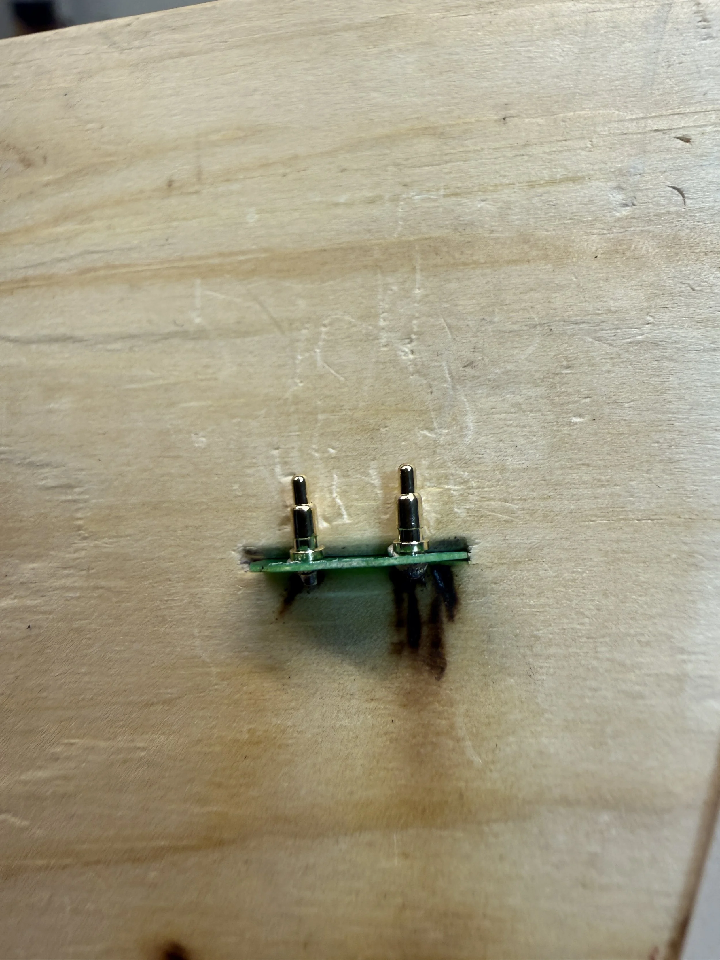 Close-up of a small green circuit board with two metal pins, mounted on a light wooden surface, with some burned marks underneath.