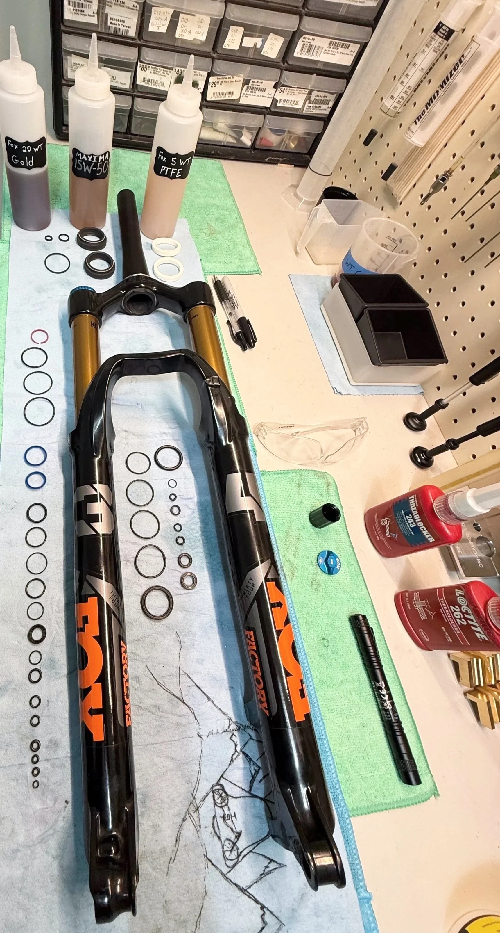 Disassembled mountain bike suspension fork and various seals and O-rings on a workbench, with oils and tools nearby.