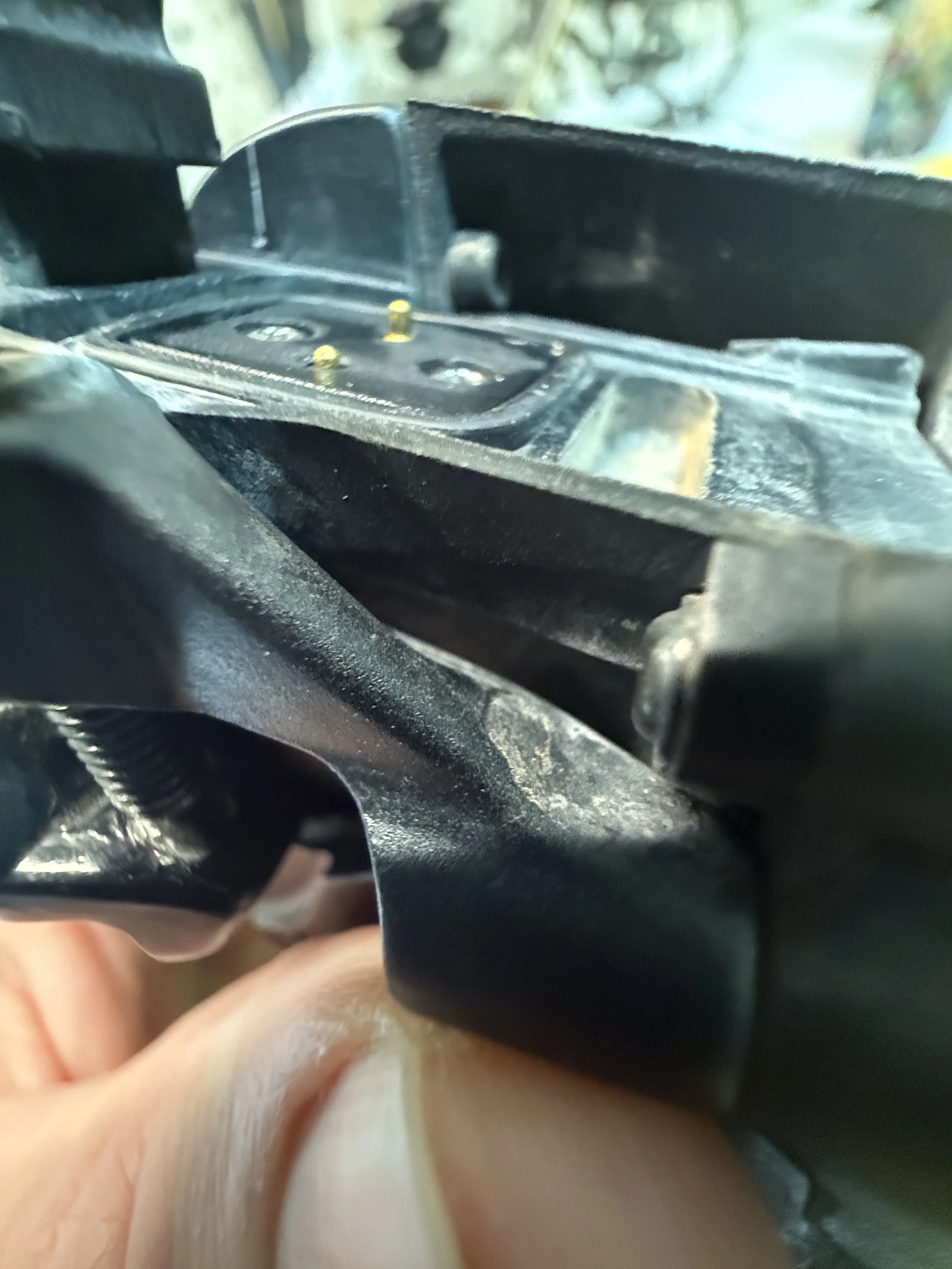 Close-up of a damaged black plastic or metal component with visible screws and metal pins, possibly part of a mechanical or electronic device.