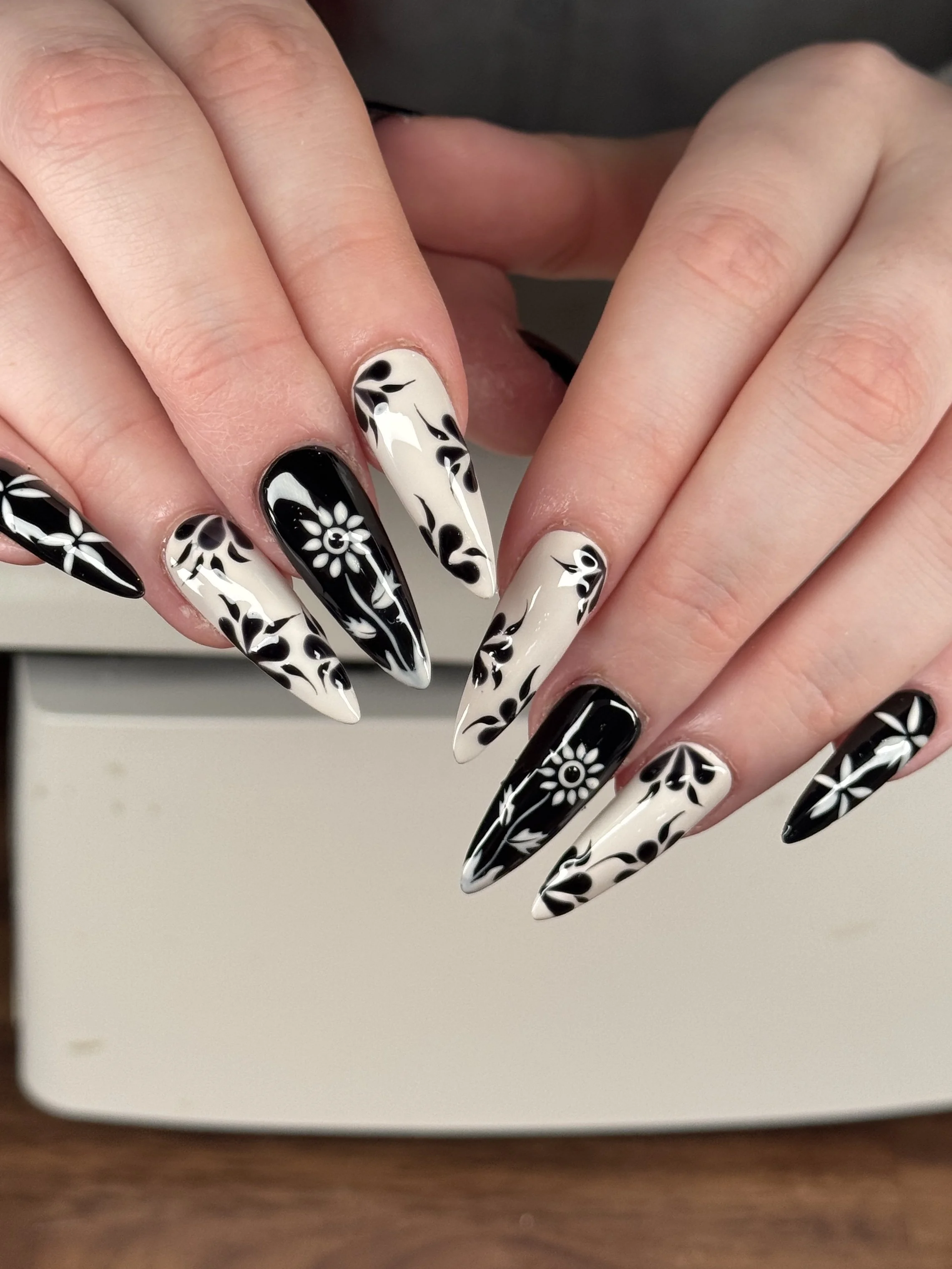 Close-up of hands with long, pointed nails painted in black and white with floral and leaf patterns.