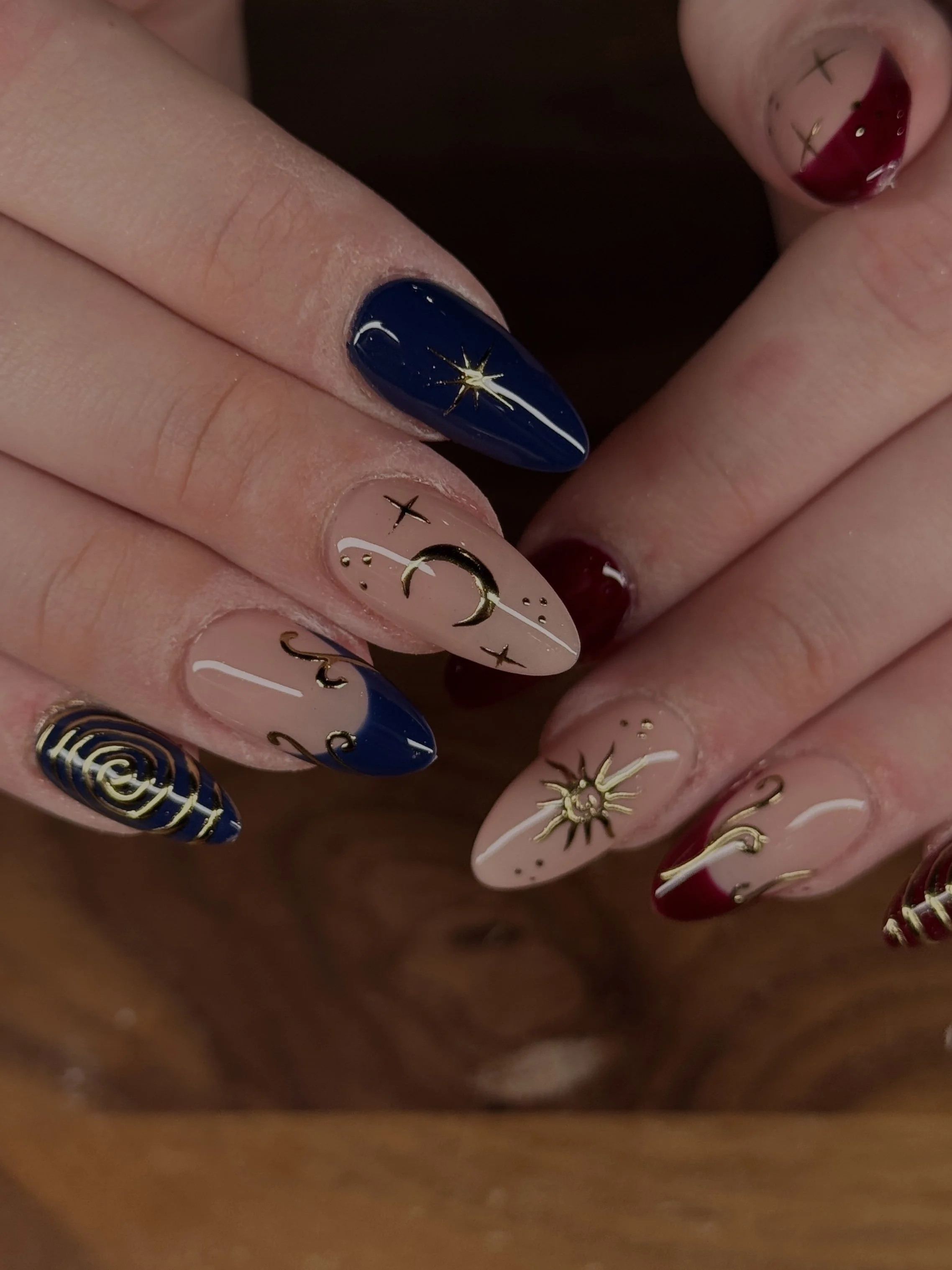 Nail art featuring celestial designs, including stars, moons, and suns, with a mix of navy blue and beige colors, accented with metallic gold details.