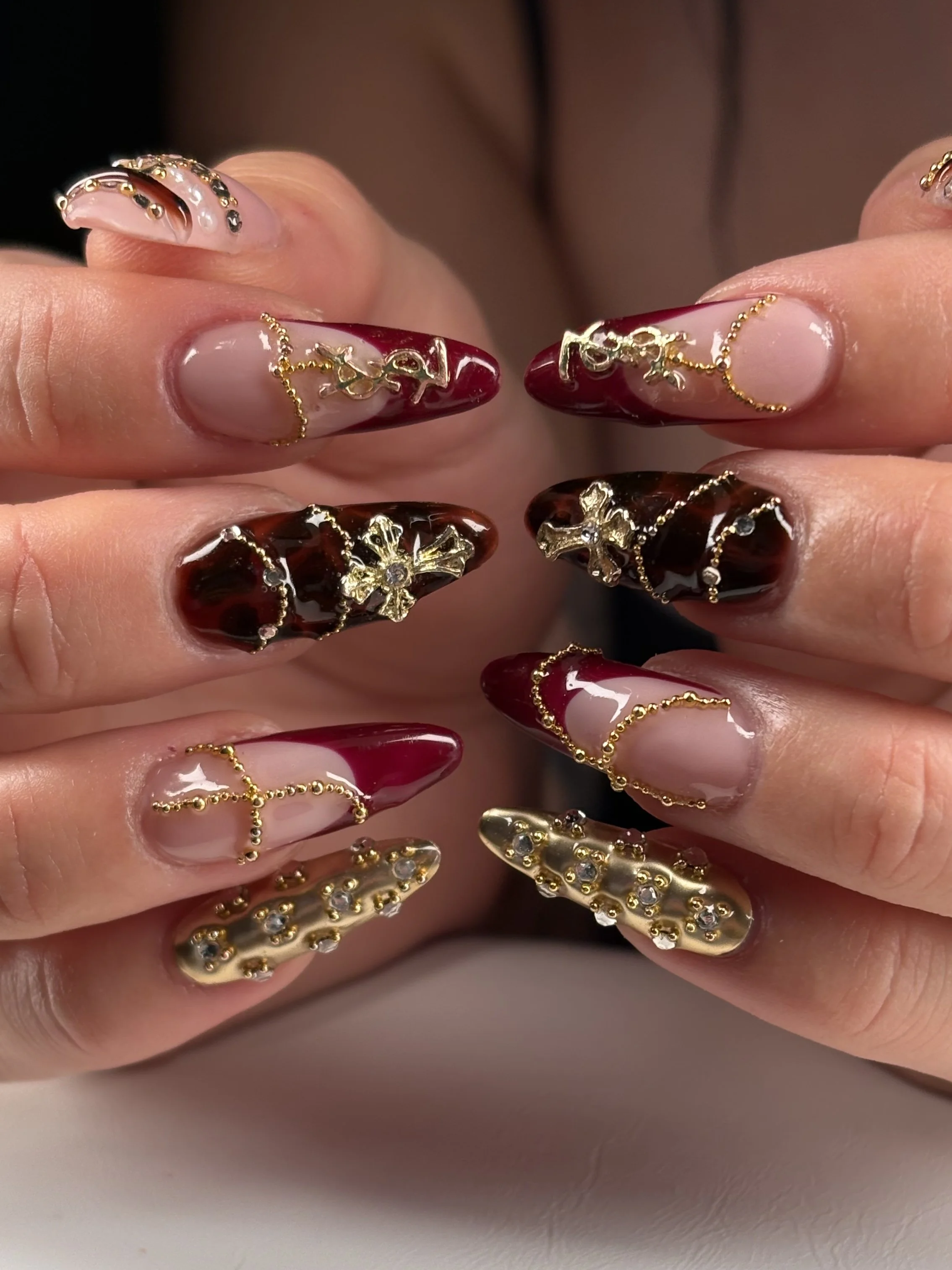 Close-up of hands with elaborately decorated nail art featuring gold chains, rhinestones, and a mix of red, black, clear, and nude colors, with a focus on ornate, long, acrylic nails.
