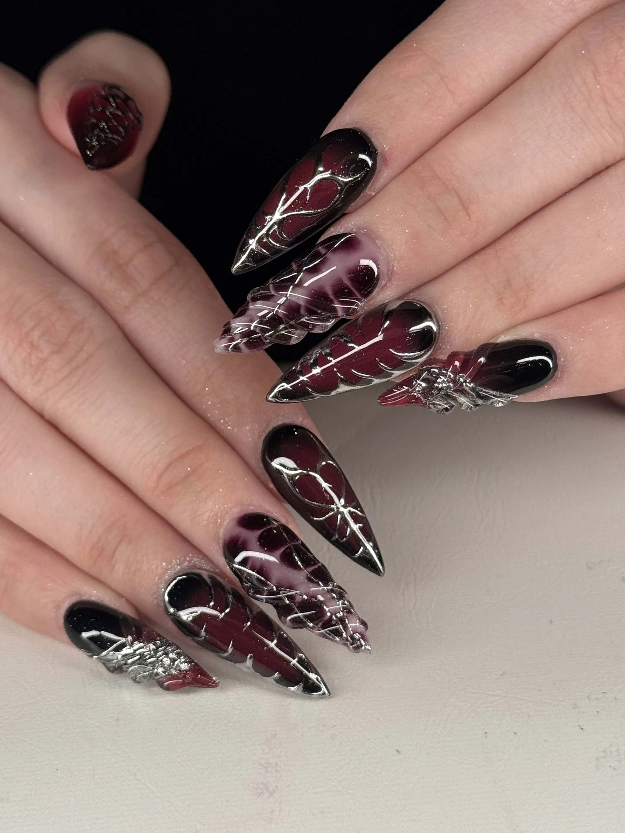 Close-up of long stiletto-shaped nails with intricate black and burgundy gradient design, silver line accents, and clear 3D embellishments.