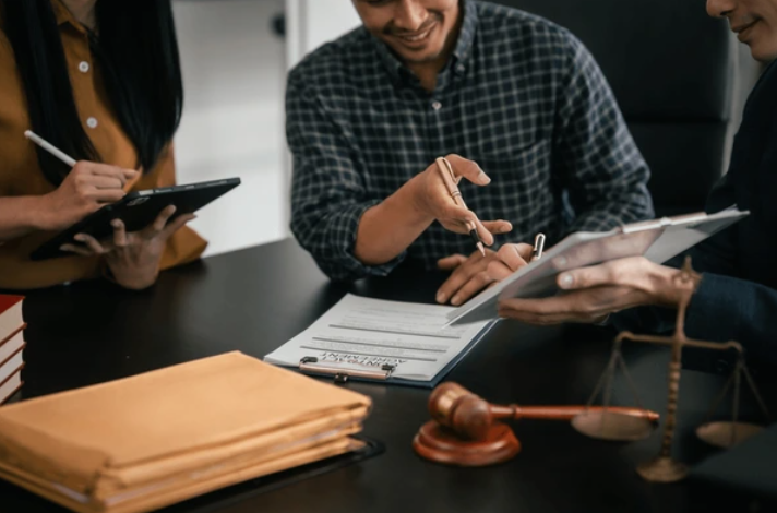 Three people at a desk with legal documents, a gavel, and scales of justice, engaged in a discussion.