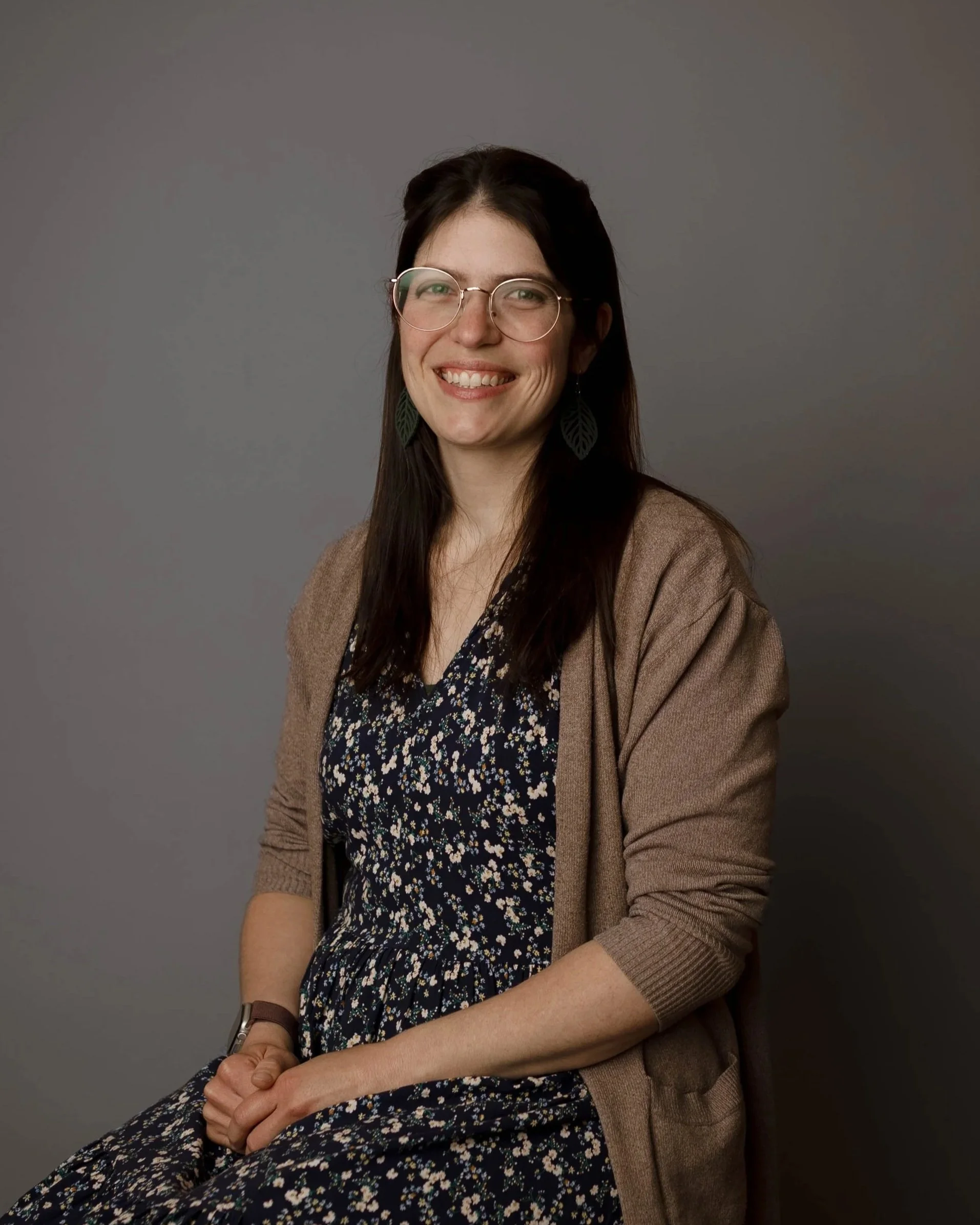 A young woman with long dark hair, wearing glasses with round frames, green leaf-shaped earrings, a floral dress, and a brown cardigan, sitting against a plain gray background, smiling with her teeth showing, hands clasped on her lap.