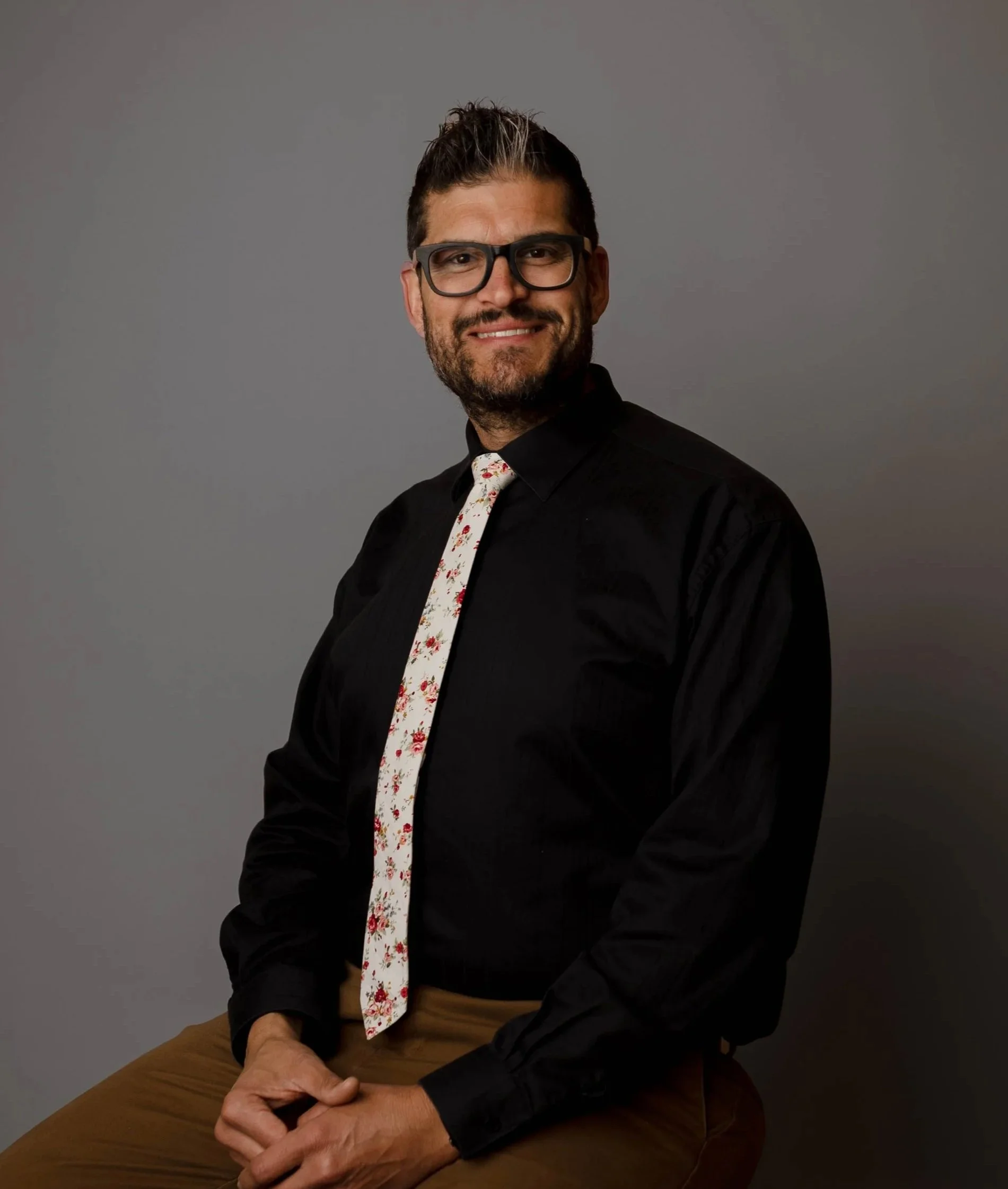 A man with glasses and a beard, wearing a black shirt and a floral tie, sitting against a grey background.