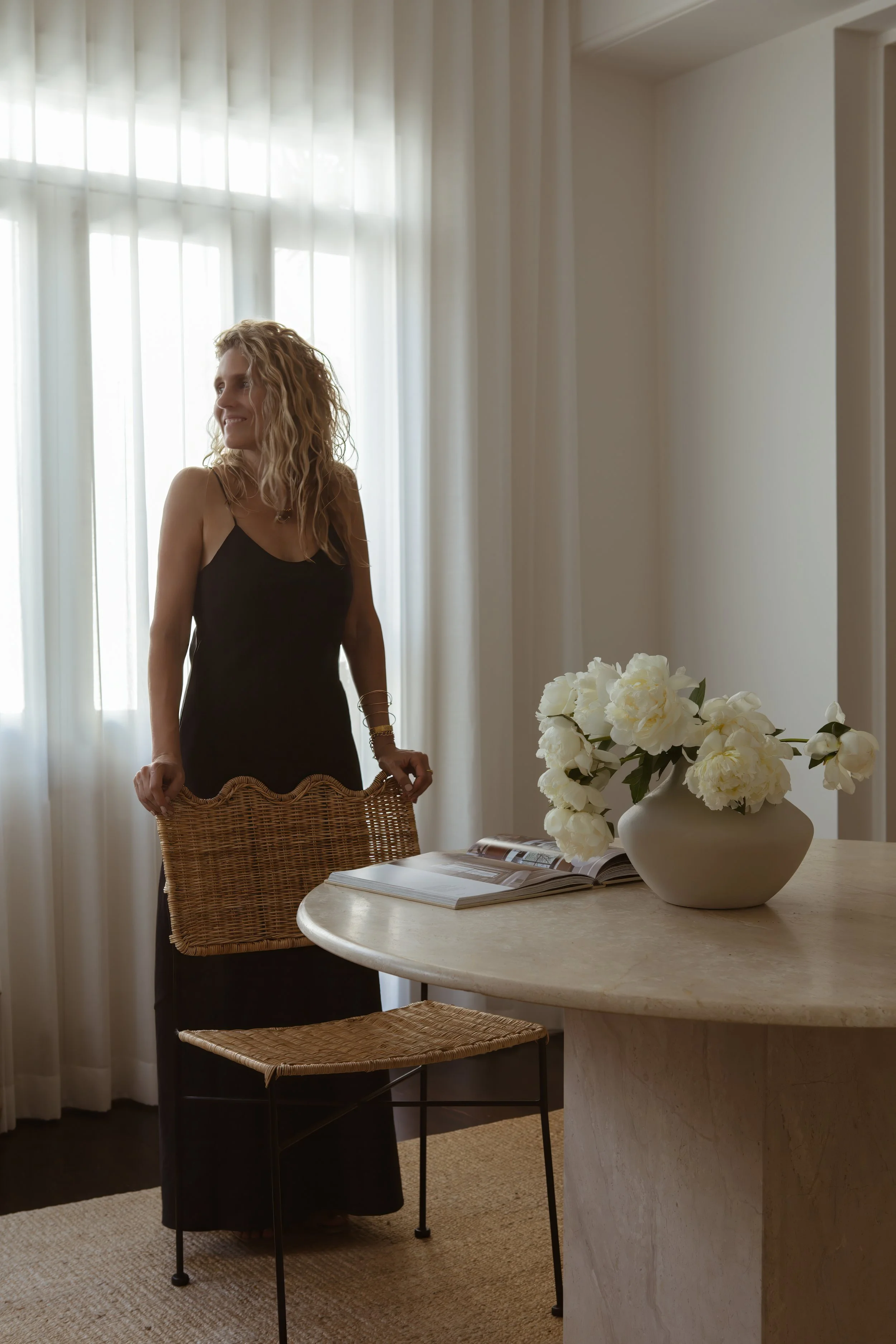 A woman in a black dress standing behind a wicker chair in a well-lit room with a large window, white curtains, a round table with a white vase of white flowers, and an open magazine.
