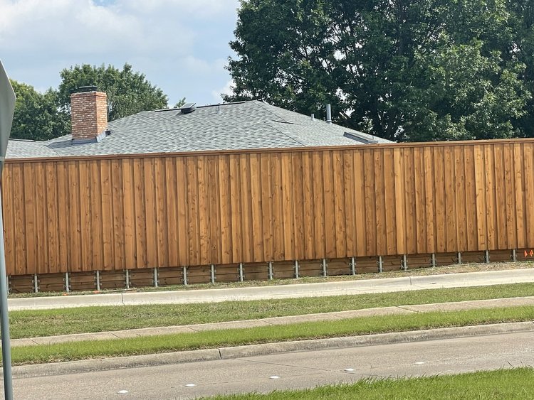 Wooden privacy fence with single-story house and chimney behind it, surrounded by trees and a lawn.