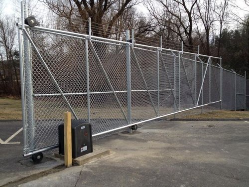 Chain-link sliding gate with security features, set in an outdoor area on a concrete surface surrounded by trees.