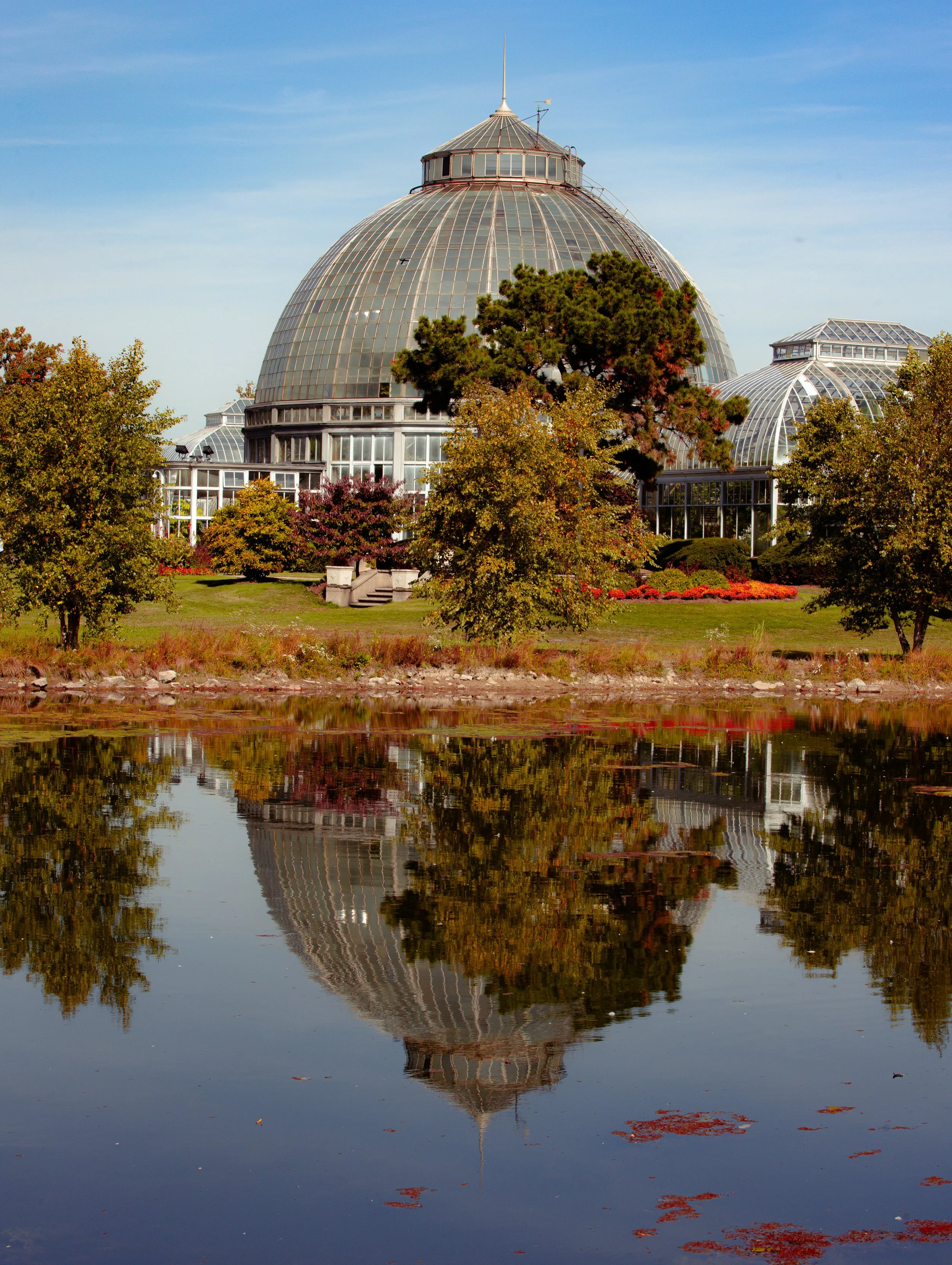 The botanical garden conservatory, or known as Belle Isle, with a glass dome, is surrounded by trees and reflected in a lake below.