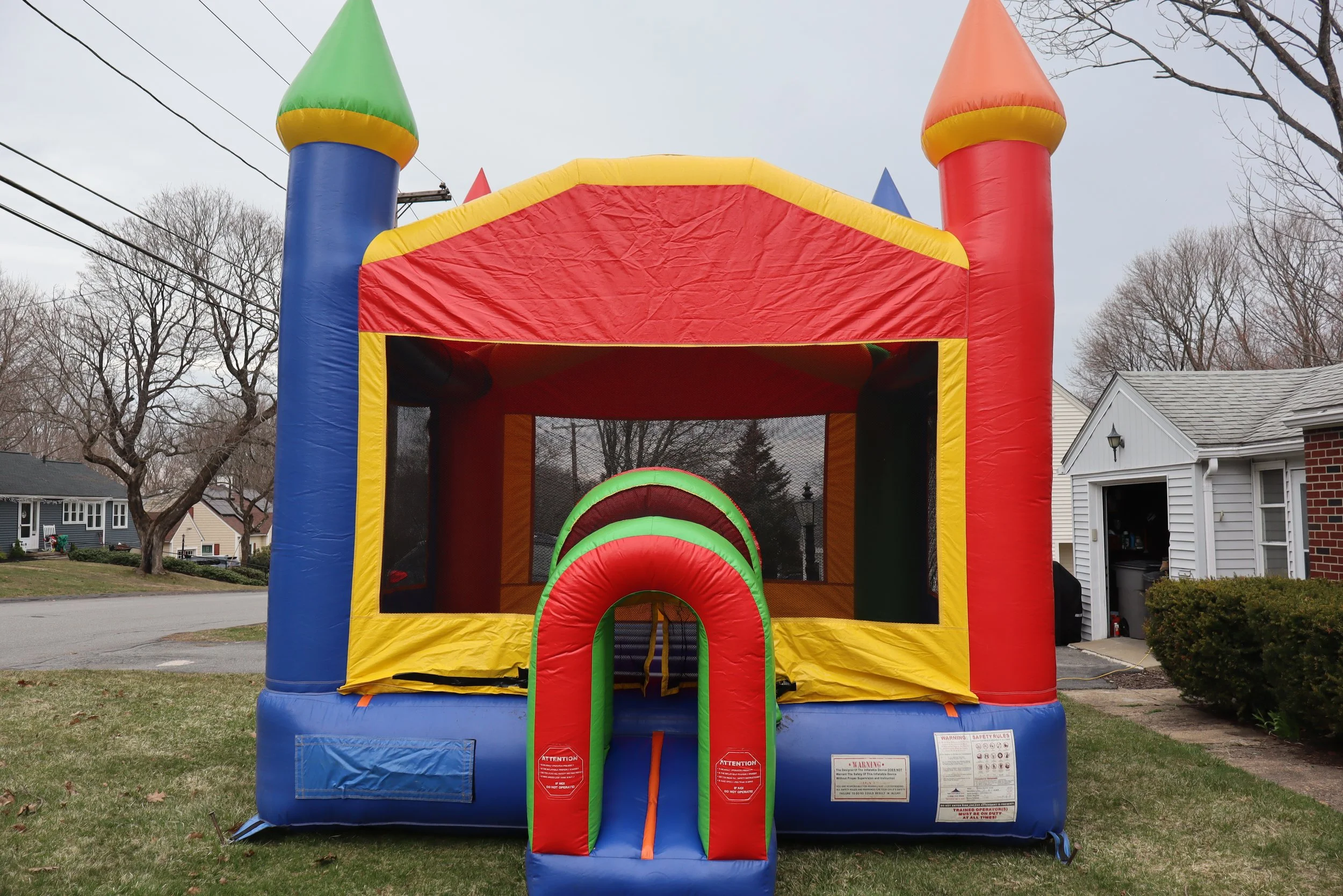 Colorful inflatable bounce house designed to look like a castle, with red, yellow, green, and blue sections, set up outdoors on a grassy area near residential houses on a cloudy day.