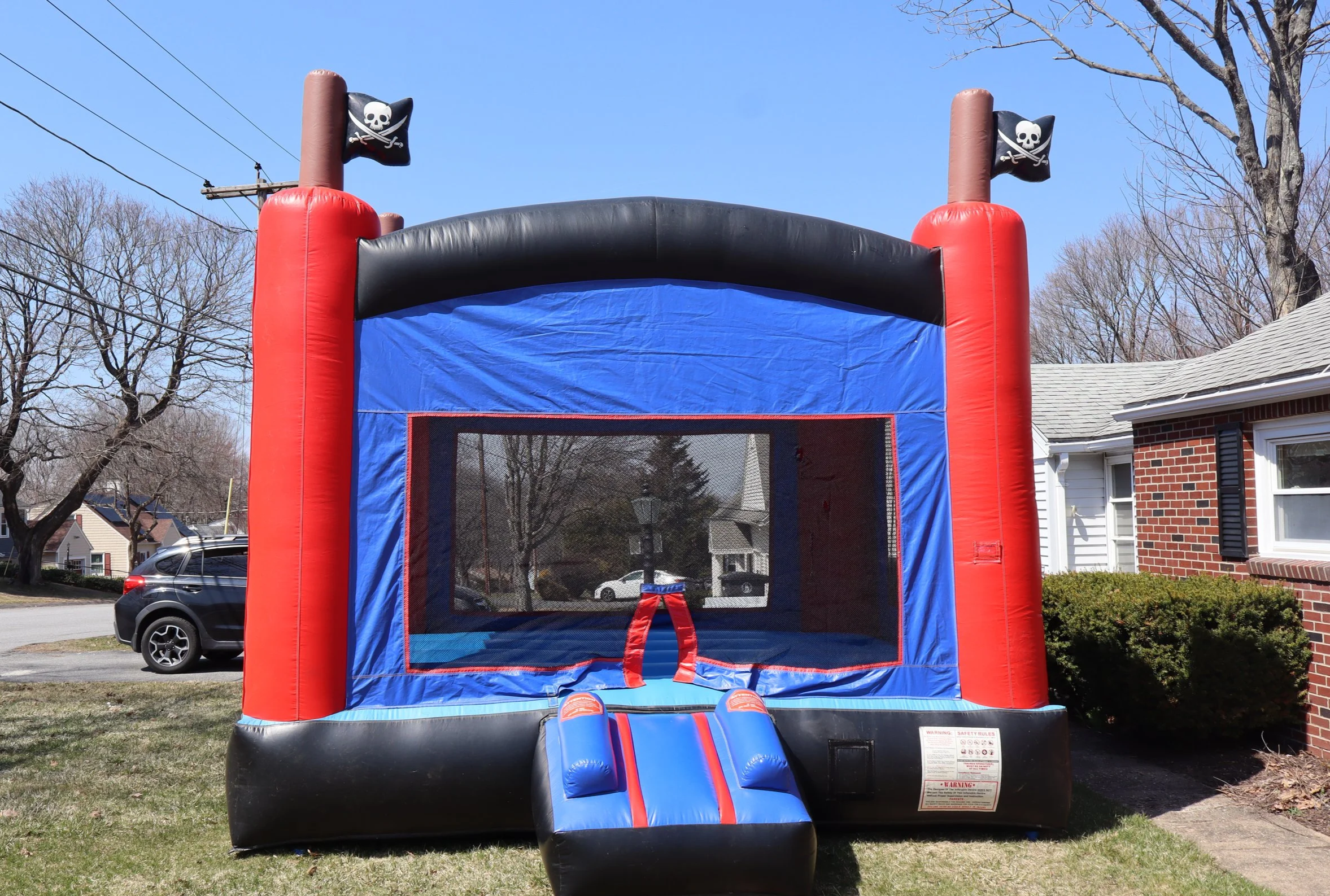Blue and black inflatable bounce house designed with pirate flags, situated outdoors on a grassy area in front of a house and other residential buildings.