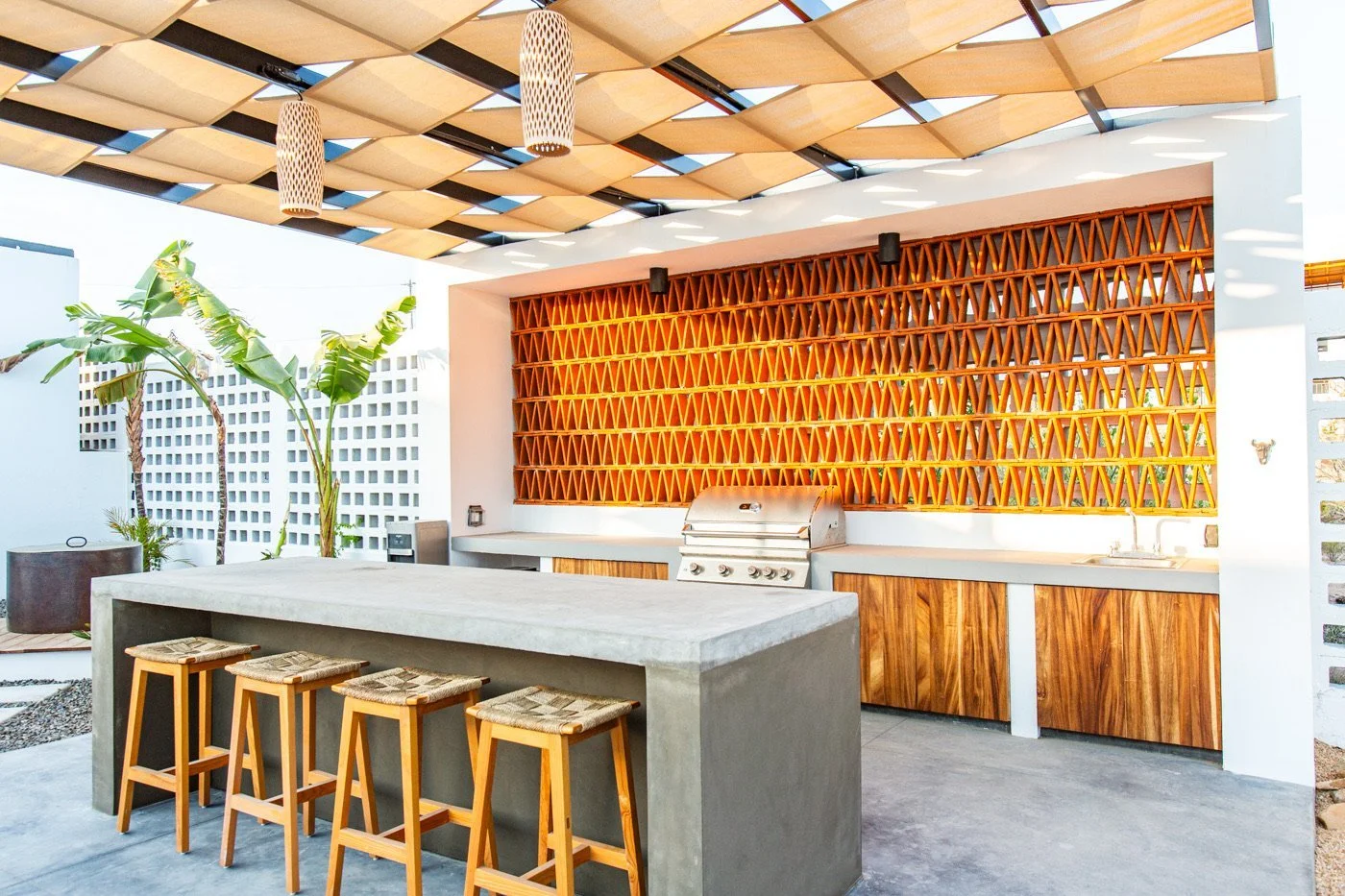 Outdoor kitchen with a concrete island, four wooden bar stools, a grill, wood cabinet doors, and decorative wooden lattice wall, with a patterned wooden roof and tropical plants.