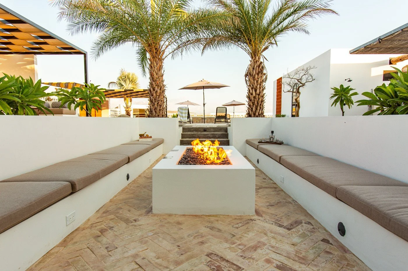 Outdoor lounge area with two long built-in benches with beige cushions on white walls, a modern rectangular fire pit in the center, and palm trees in the background. There are sun umbrellas, lounge chairs, and a clear sky.