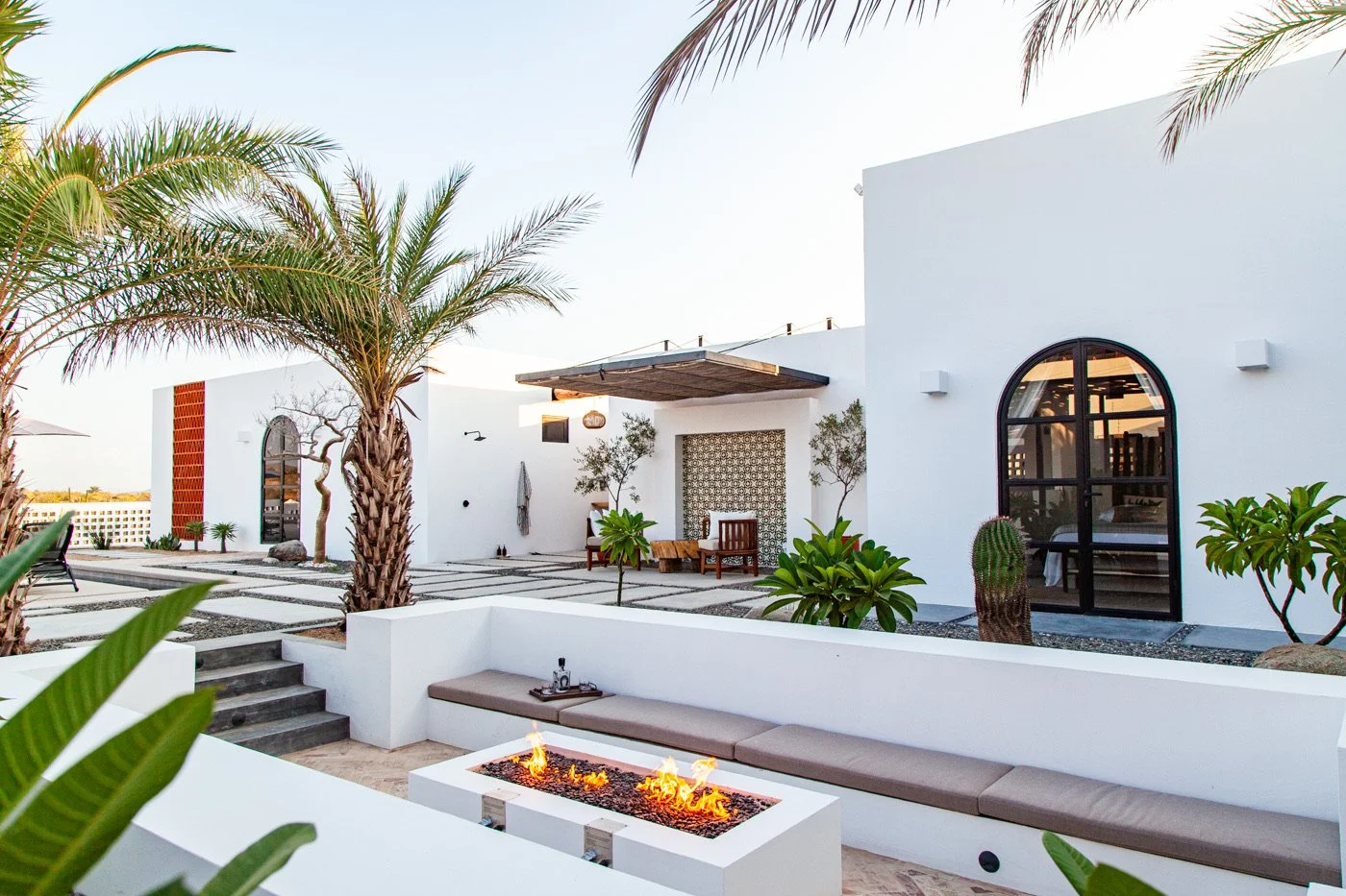 Modern white house with outdoor seating area, palm trees, and a fire pit, in a sunny setting with clear sky.