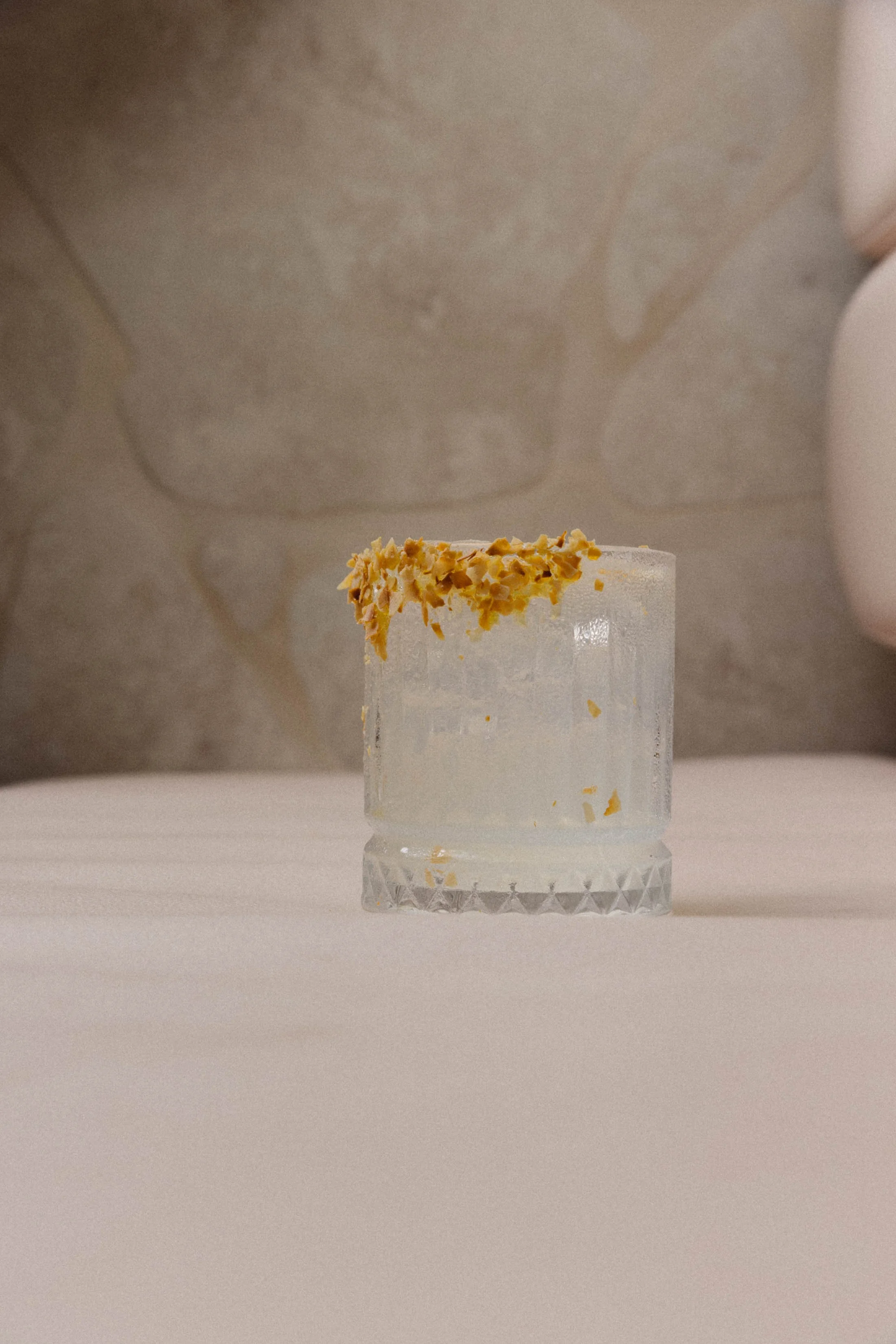 A glass with a textured surface partially filled with a clear beverage. The rim of the glass is adorned with crushed nuts or granules. The background is a neutral-toned wall and surface.