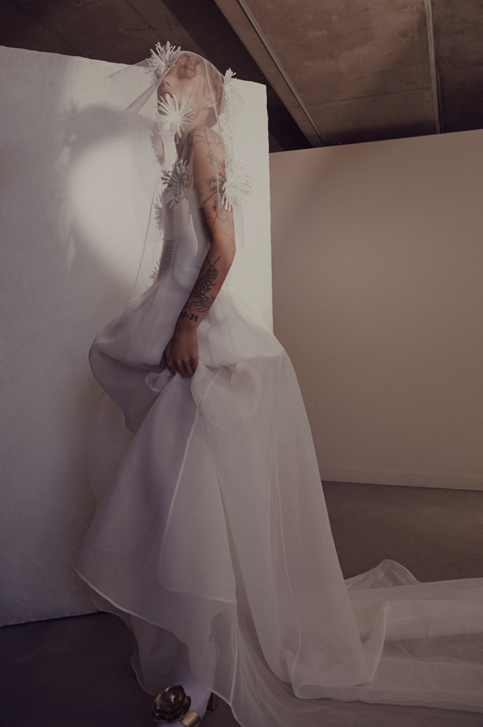 Person in a flowing white gown with floral veil and tattoos, standing indoors.