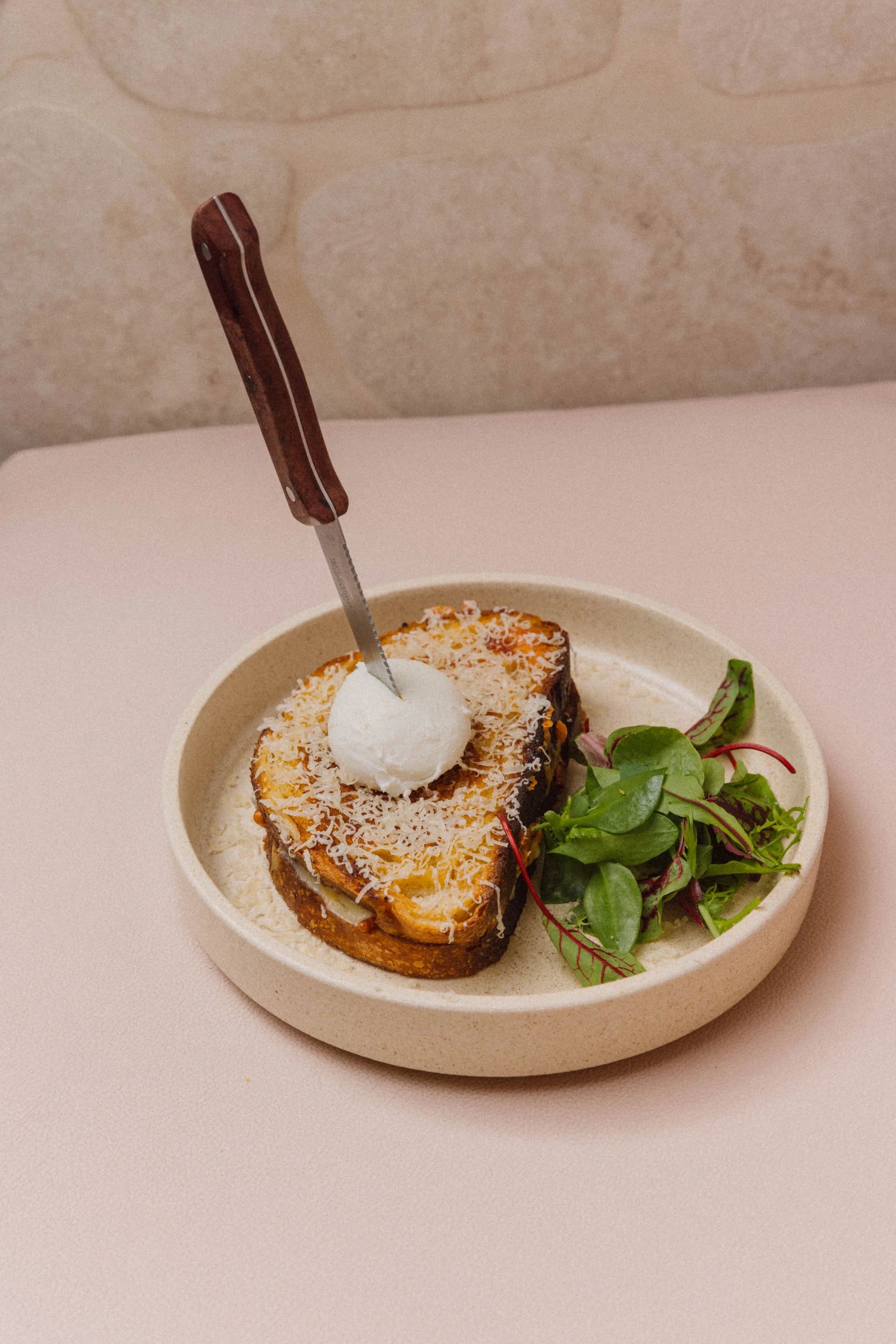 Plate of savory French toast topped with shredded cheese and a poached egg, garnished with fresh greens, with a knife inserted in the top.
