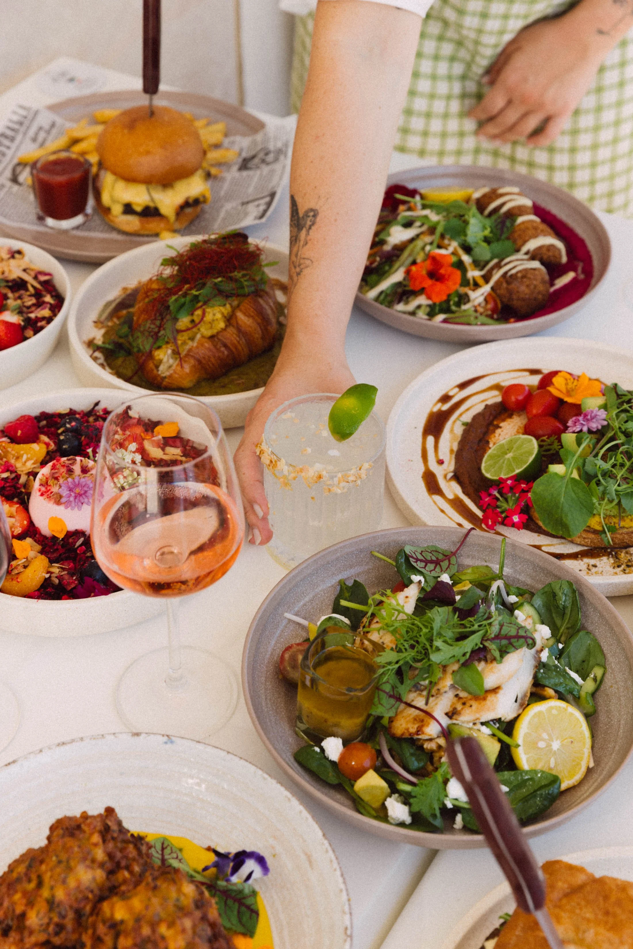 Assorted dishes on a table including a burger with fries, salads with greens and tomatoes, a pink dessert with fruits and flowers, beverages including a rosé wine and a cocktail with a lime wedge, and a person reaching for a drink.
