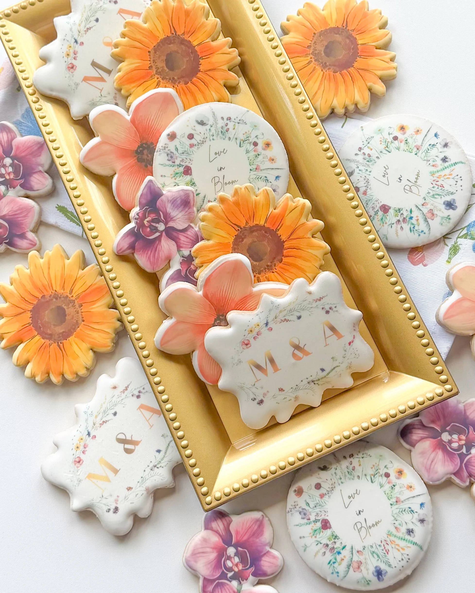 Decorative cookies shaped like sunflowers, orchids, and circles with floral designs and the phrase 'Love in Bloom' on some.