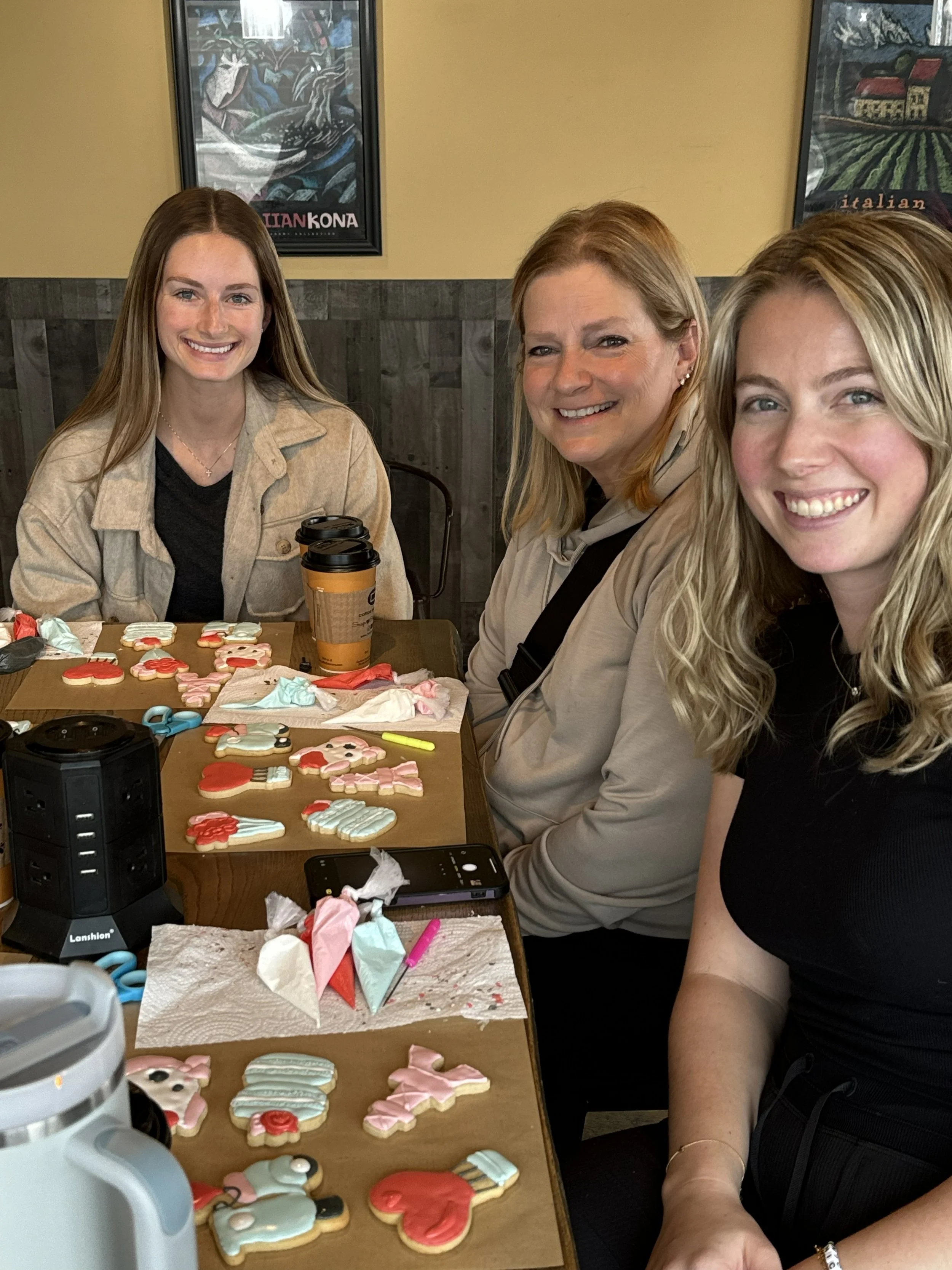 Three women sitting at a table decorated with various decorated cookies in holiday shapes, with baking supplies and coffee cups, smiling at the camera in a cozy indoor setting.