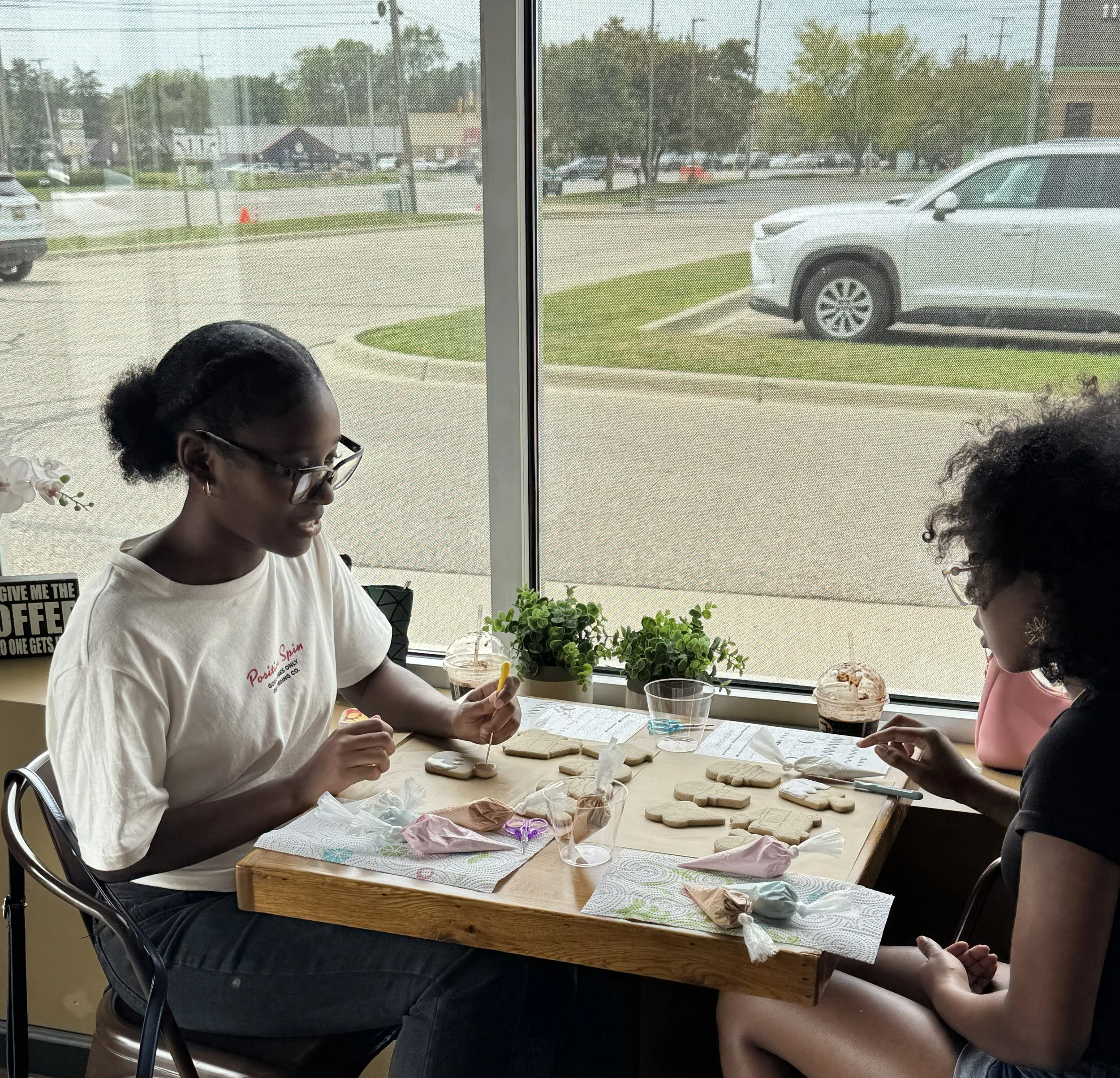 Two women with curly hair and glasses sit at a wooden table by a large window, decorating cookies with frosting and sprinkles.