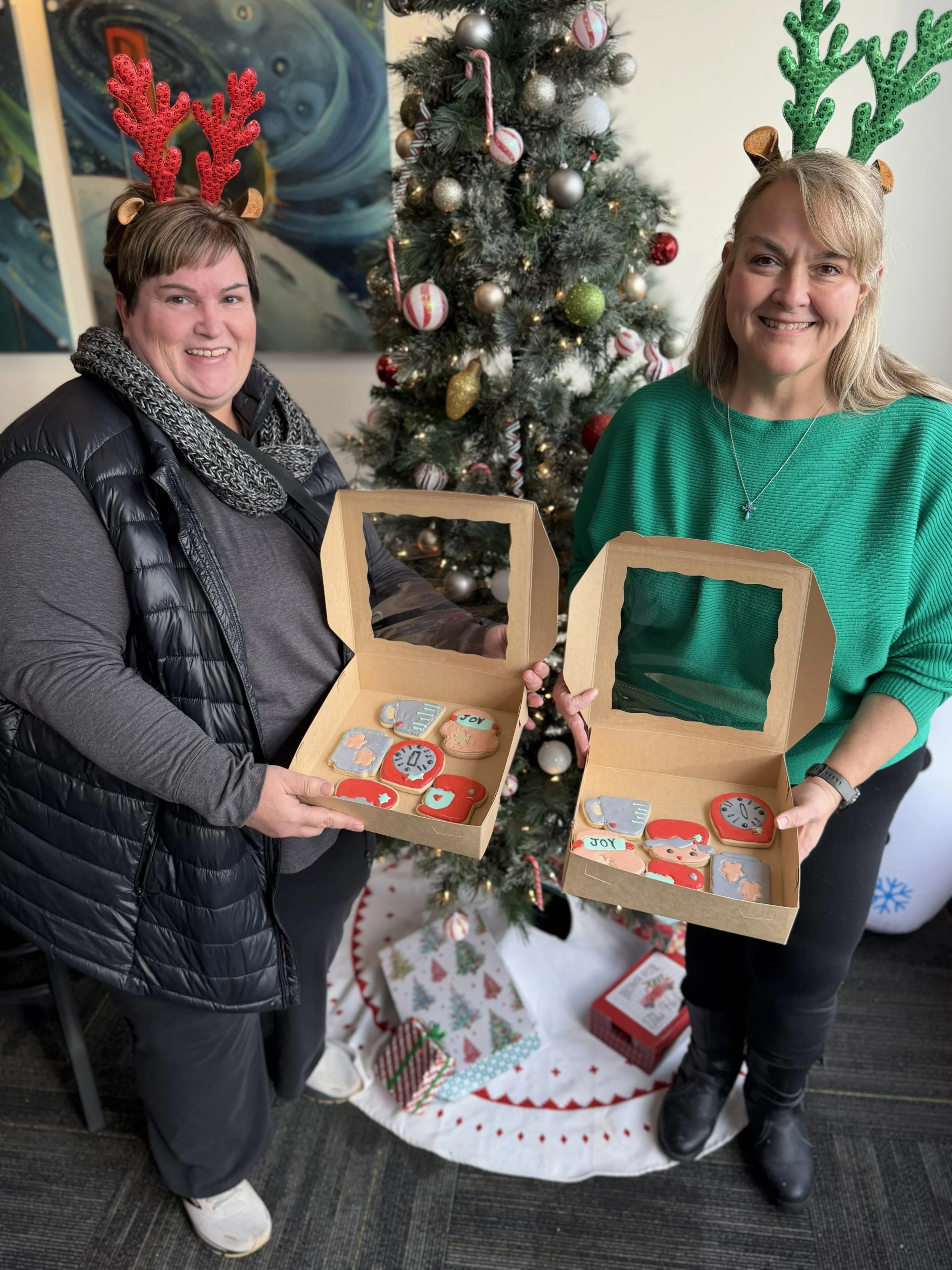 Two women smiling and holding open gift boxes with decorated cookies, standing in front of a Christmas tree with ornaments, candy canes, and festive decorations.