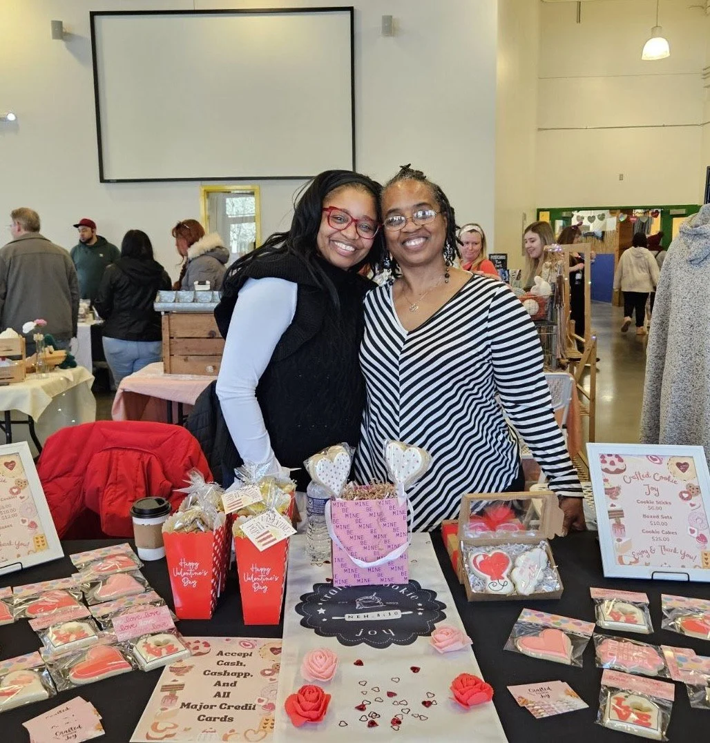 Two women smiling at a Valentine's Day-themed craft stall at an indoor market event, with various cookies and decorations on display.
