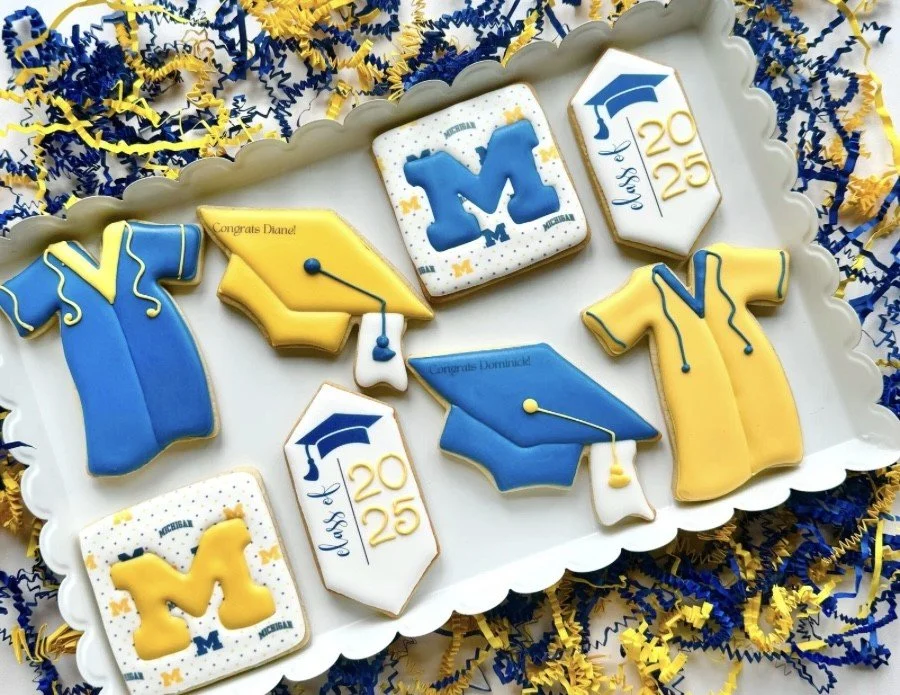 Assorted University of Michigan graduation-themed cookies with blue and yellow colors, including caps, gowns, and school logo, on a white tray surrounded by blue and yellow confetti and streamers.