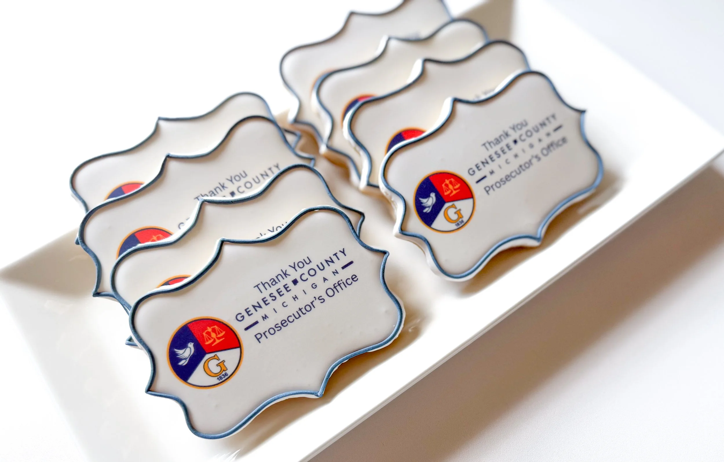 A tray of eight decorative cookies with a scalloped edge, featuring a logo with a scale and a bird, and the words "Thank you GENESEE COUNTY MICHIGAN Prosecutor's Office".