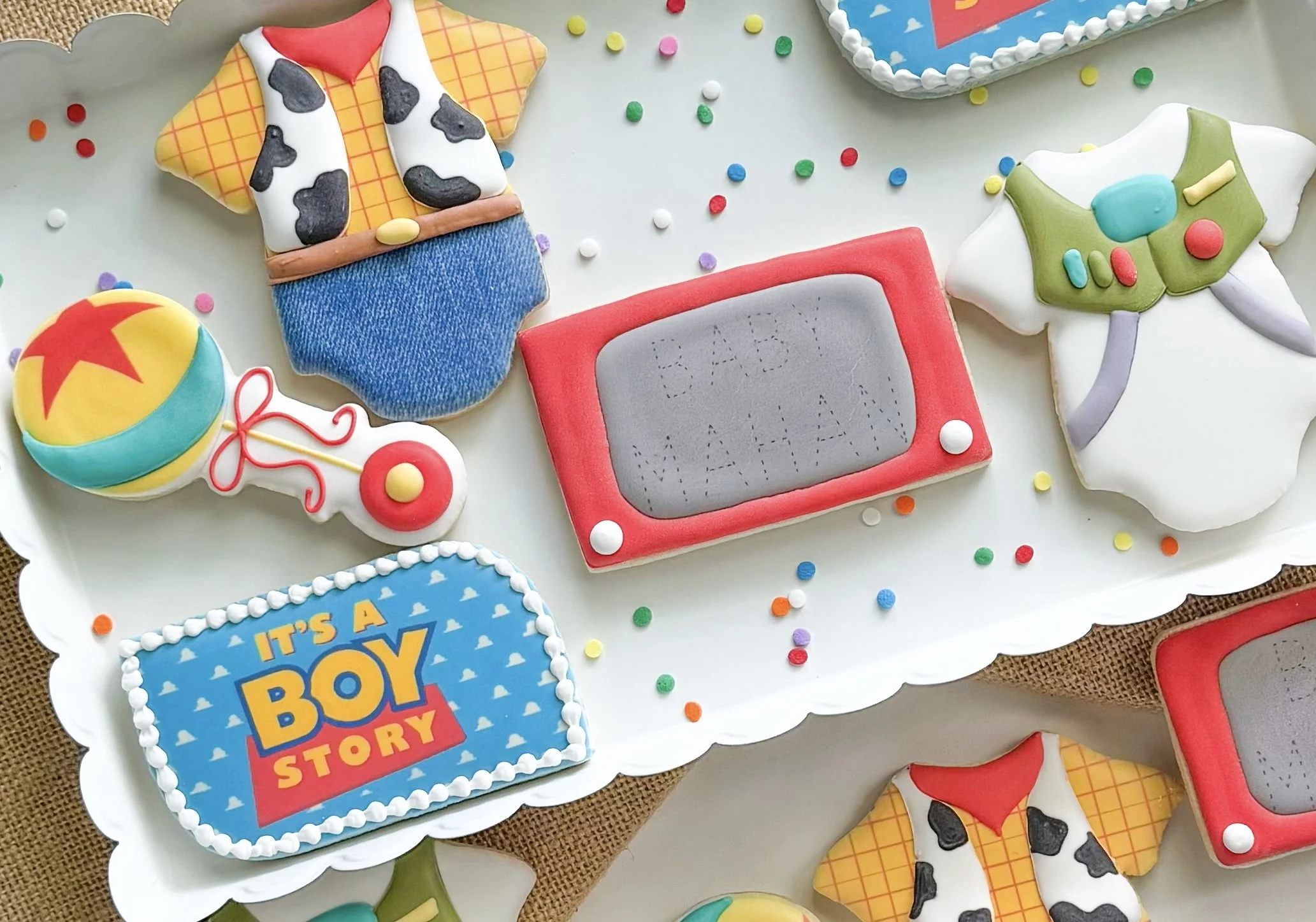 Decorated cookies themed for a toy story baby shower, including a cowboy onesie, a Buzz Lightyear themed onesie, a red etch-a-sketch, a blue ball, and a "Boy Story" plague cookie.