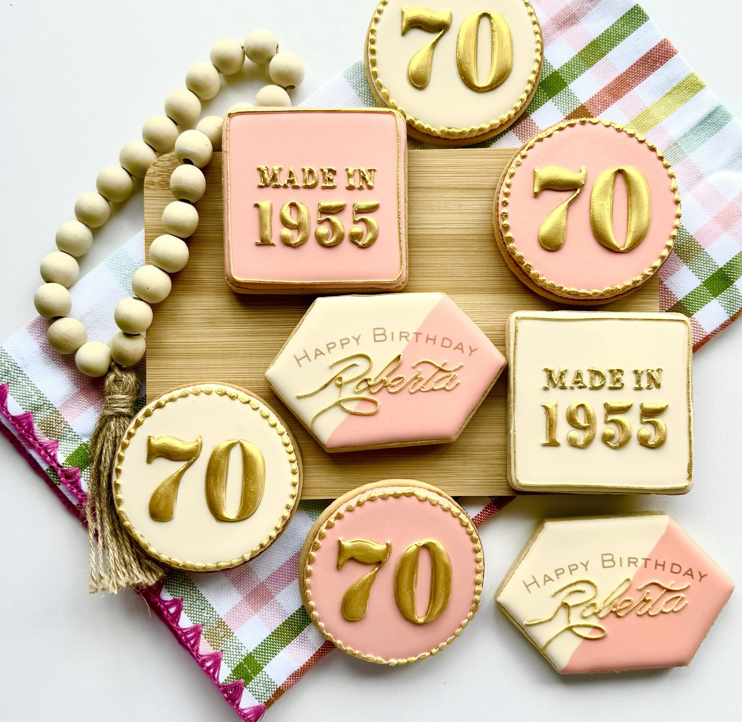 Decorative cookies celebrating a 70th birthday, some with the number 70 in gold, some with 'Made in 1955', and others with 'Happy Birthday Roberta' in elegant script, arranged on a wooden board with a pink and green plaid cloth.