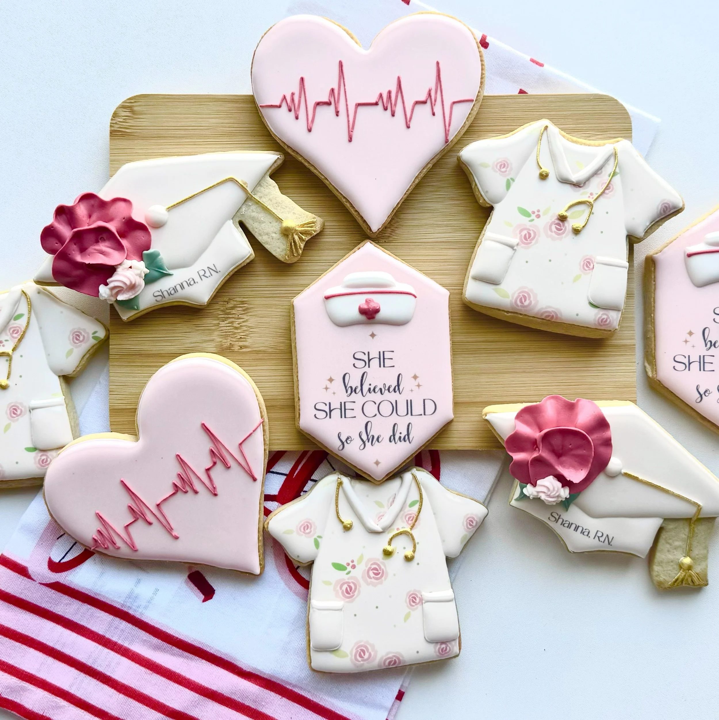 Decorative medical-themed cookies on a wooden board, including hearts with heartbeat lines, nursing scrubs, stethoscopes, and a trauma bag, with a quote that reads 'She believed she could so she did', all themed around healthcare and nursing.
