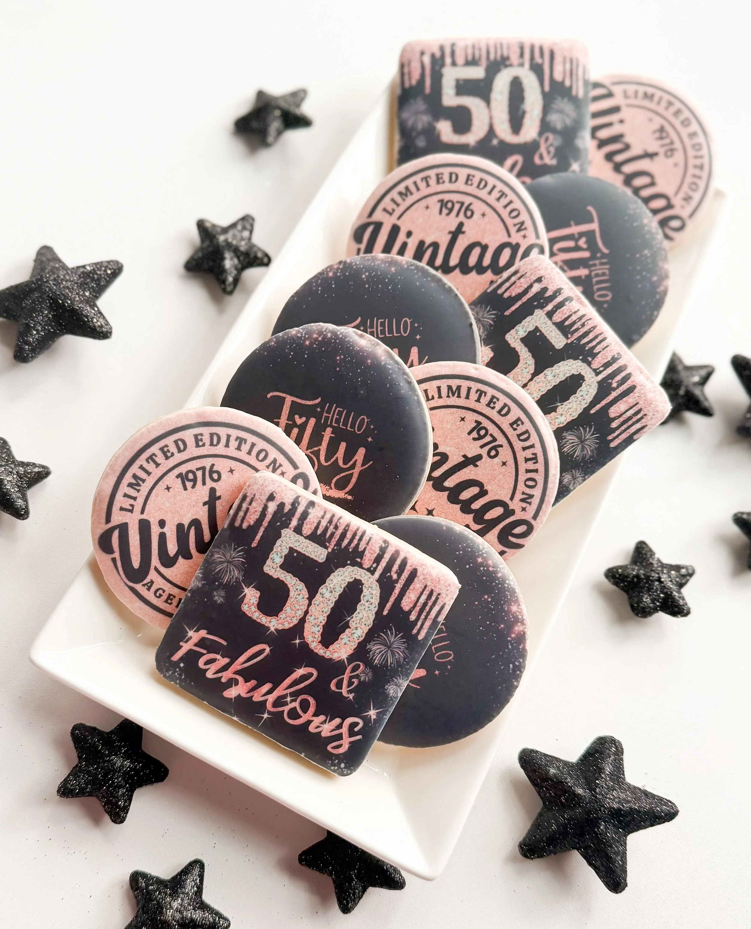A white plate with several decorative cookies featuring pink and black designs and text celebrating a 50th anniversary, with black star-shaped glittery decor scattered around.