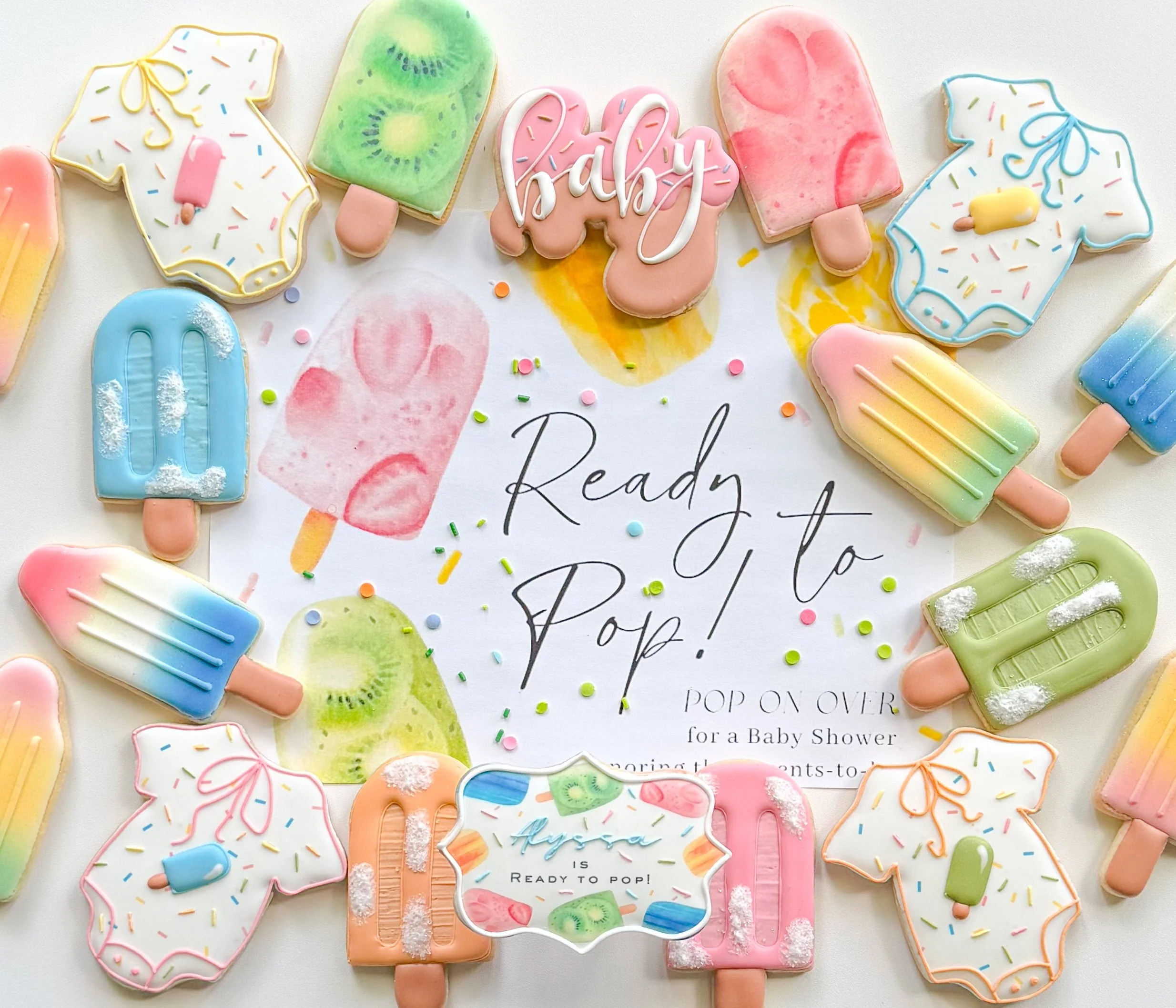 Colorful cookies shaped like popsicles and baby onesies, decorated with pastel icing and sprinkles, arranged around a baby shower card.