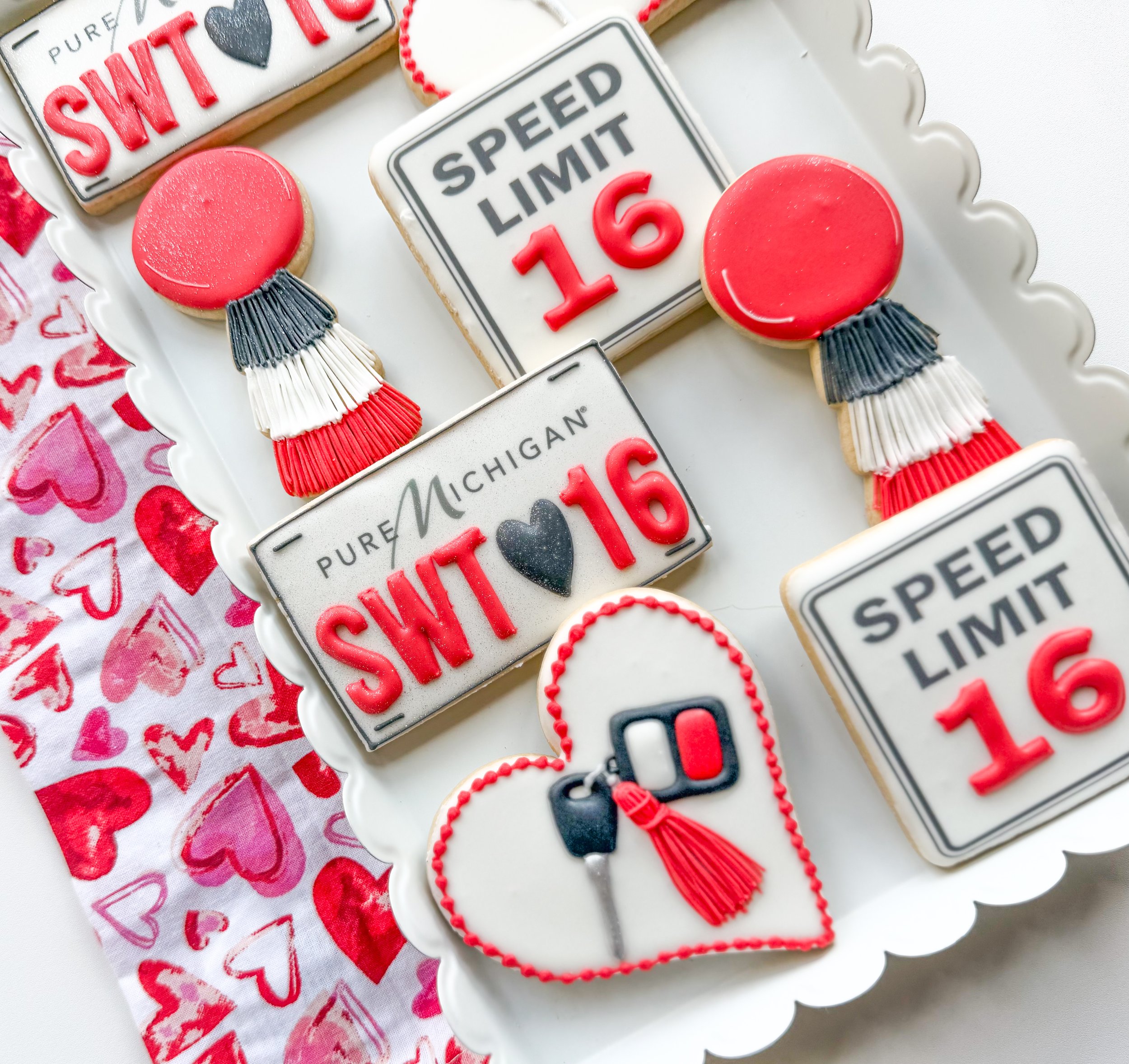 Decorative cookies with Sweet 16 themes, including a license plate style cookie reading 'Speed Limit 16,' a red and white heart shaped cookie, and a balloon shaped cookie with fringe, all arranged on a jewelry white scalloped dish.