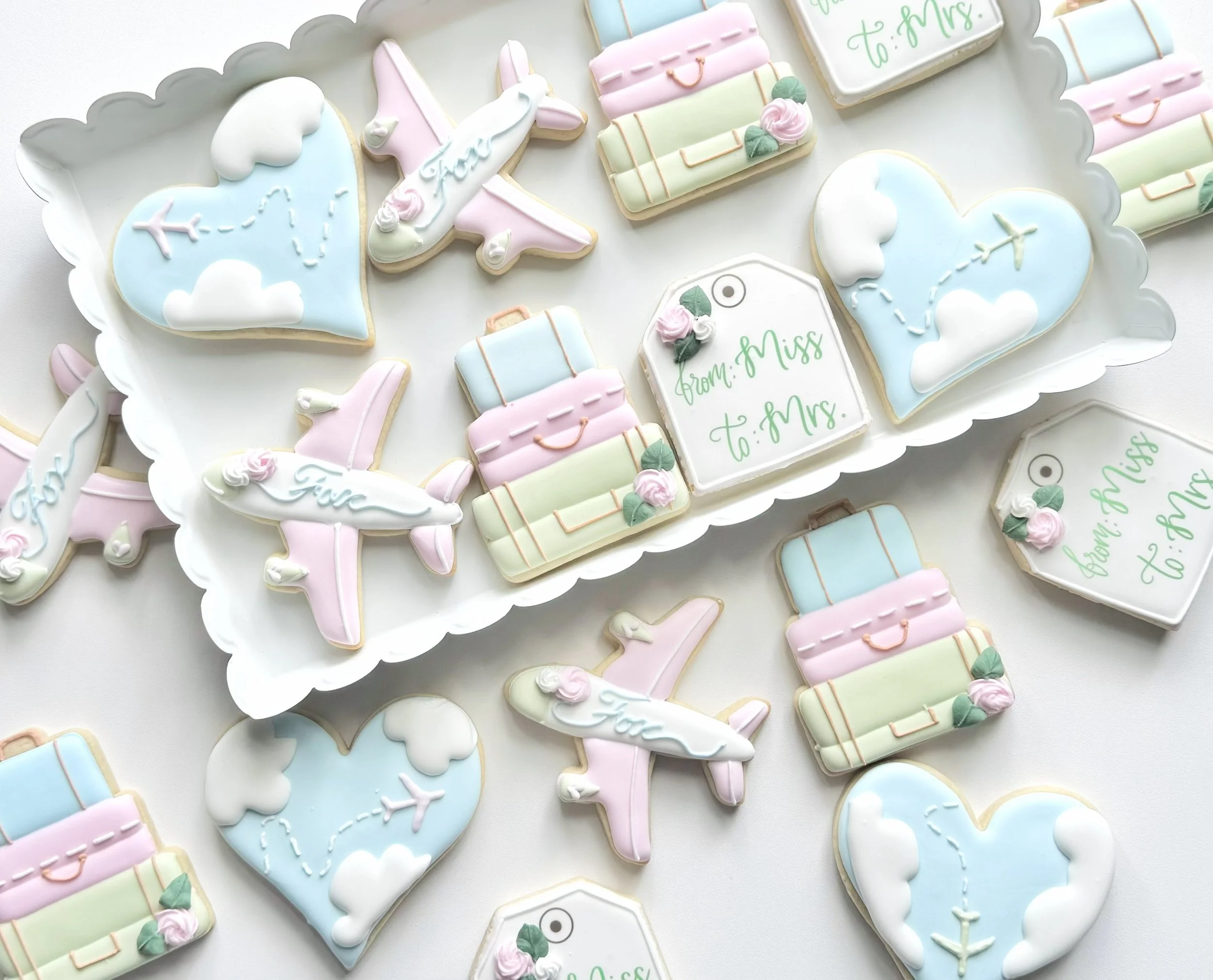 Decorative cookies themed for travel or a honeymoon, including airplanes, hearts with cloud and plane designs, suitcases, and luggage tags, on a white surface.
