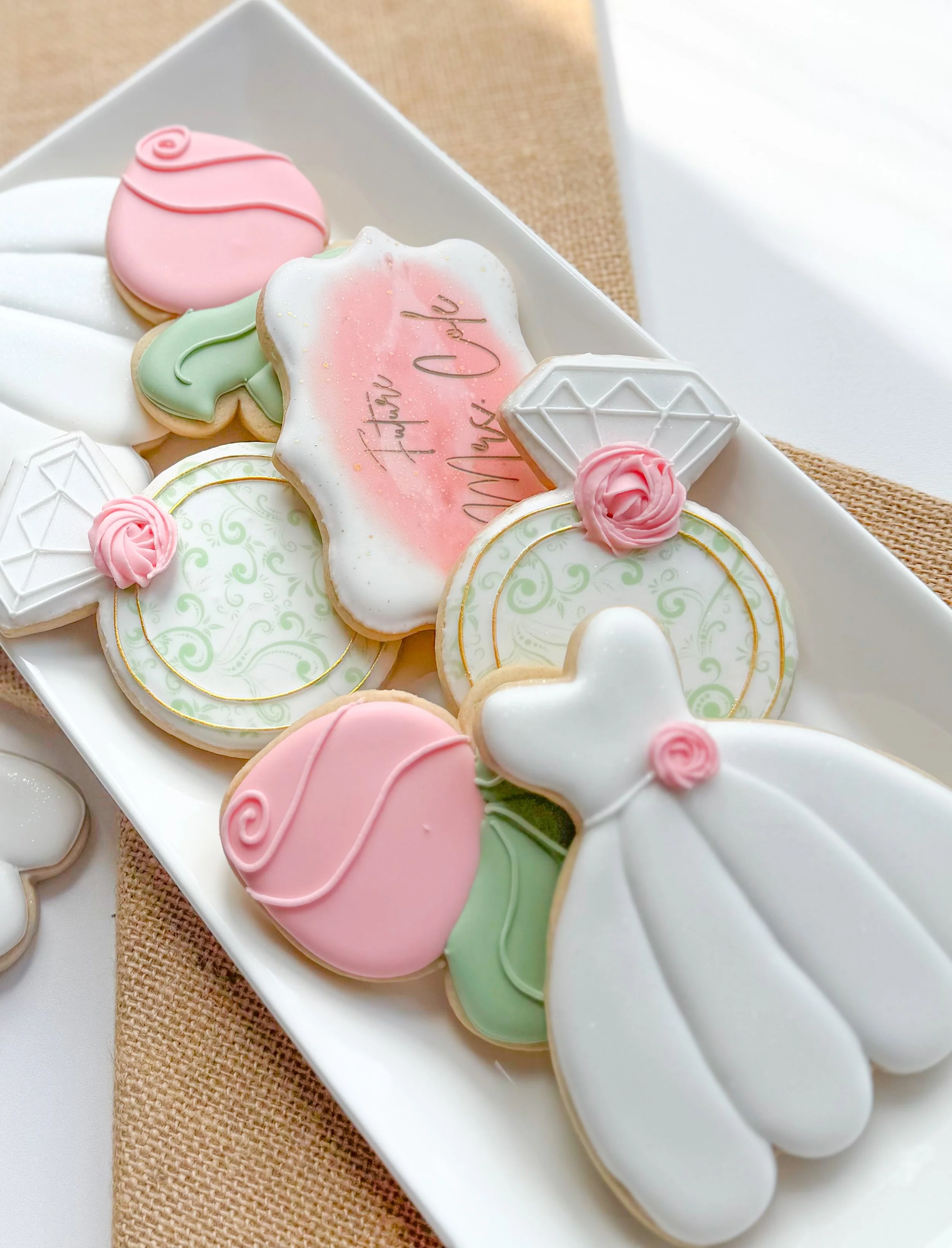 Assorted decorated cookies on a white platter, with pink, green, and white icing, some with gold accents, and one with a message 'Future Mrs.' written in pink and gold, on a woven cloth background.